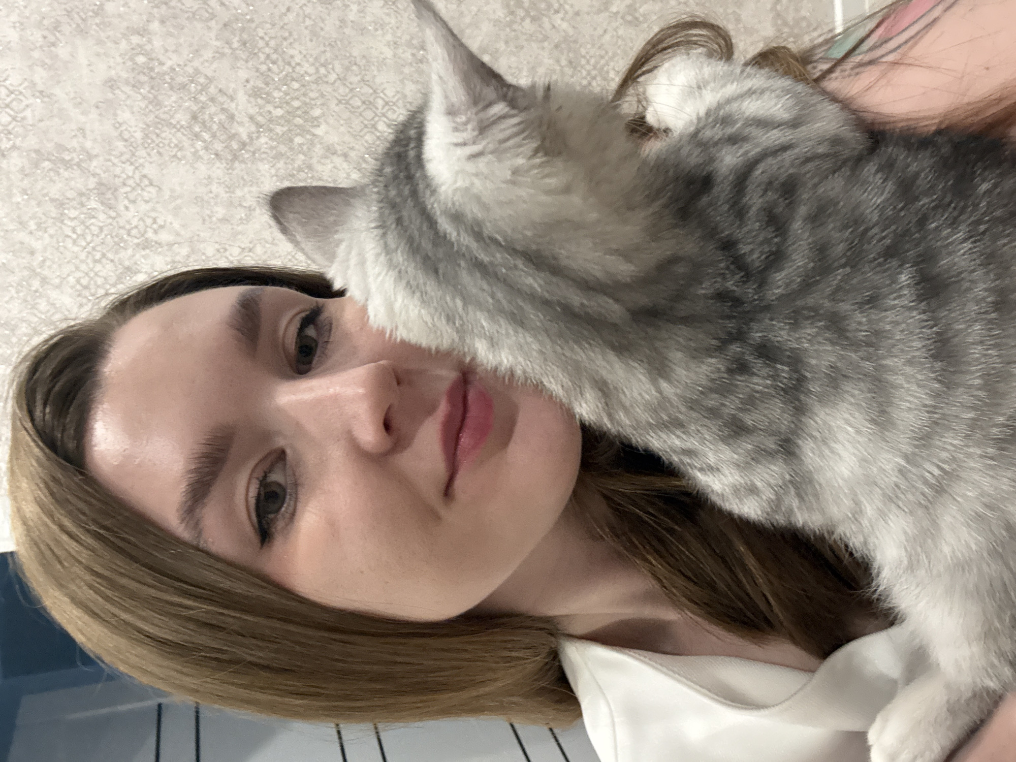 A young woman is holding a gray tabby cat close to her face. The woman has fair skin, light brown hair, and is wearing a white shirt. The background appears to be an interior space with textured wallpaper. The image is taken from a close-up perspective, focusing on the interaction between the woman and the cat. There is no specific information in the image to indicate that the location is Izhevsk, Russia.