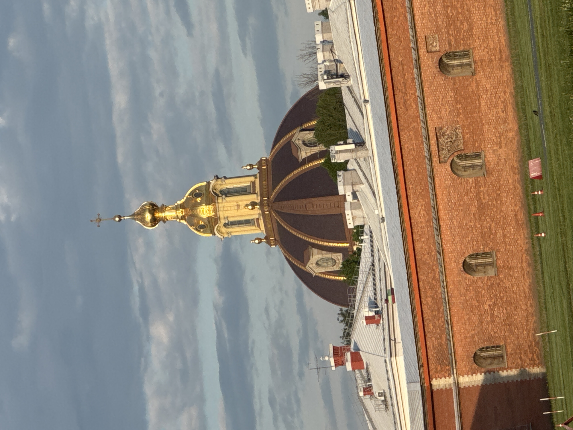 The image displays an elevated view of a prominent architectural landmark in Saint Petersburg, Russia, likely captured from a nearby rooftop. At the center-left, the ornate, gilded dome of a church or cathedral is the focal point. This dome features a large, dark brown or bronze base adorned with intricate golden vertical stripes and small round windows, surmounted by a brilliant golden onion dome topped with an Orthodox cross. The golden surfaces gleam brightly under direct sunlight, suggesting either a late morning or afternoon setting.
In the immediate foreground and extending to the right, parts of an adjacent building's flat white rooftop are visible, equipped with modern utilities such as vents and air conditioning units, creating a stark contrast with the historic dome. Further to the right, a distinctive red brick wall, possibly belonging to another building or an enclosure, features several arched window recesses and decorative rectangular inlays. Beyond this wall, a neatly maintained green grassy area stretches out. The sky above is partly cloudy, with wispy stratocumulus clouds scattered across the blue expanse. No people or discernible text are present in the scene.