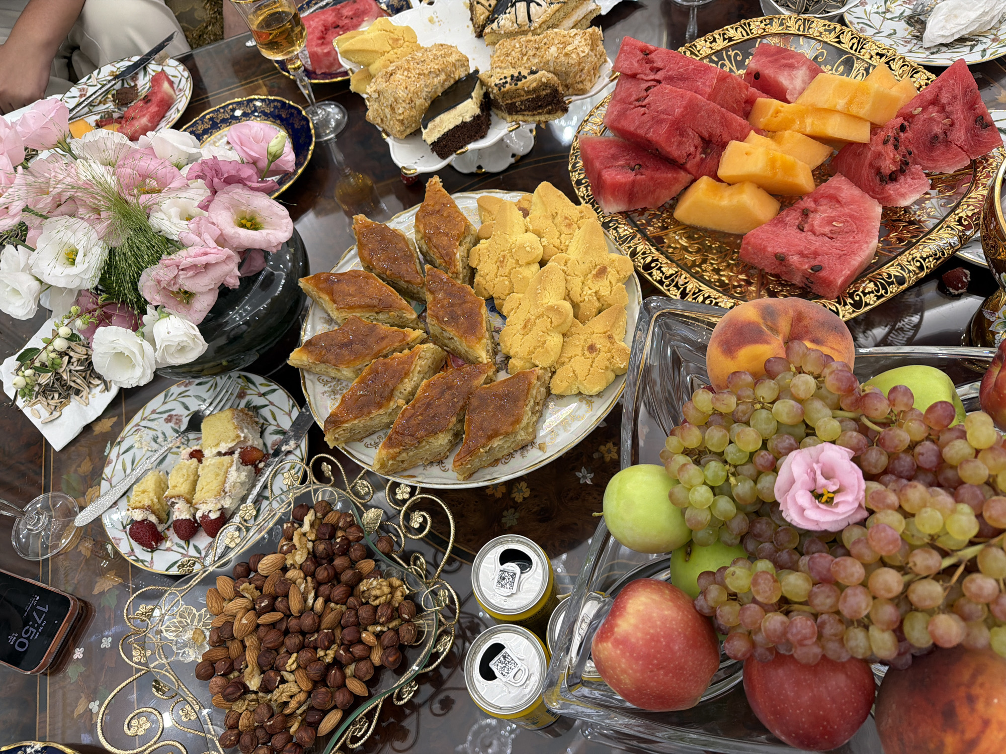 This is a close-up, overhead shot of a table laden with a variety of food and a floral arrangement. The setting appears to be an indoor dining area, possibly a home, with a dark, polished wooden table surface. The arrangement is indicative of a celebratory meal or gathering.
Dominating the center of the table is a large bouquet of pink and white flowers in a dark vase. Surrounding this are several plates of food and drinks. To the left, a clear glass bowl overflows with a mix of nuts, including almonds and walnuts. Next to this, two open aluminum cans, likely soft drinks, are visible.
A plate of pastries, including baklava and what appear to be shortbread cookies, is positioned in the lower center. To the right, a decorative gold tray holds sliced watermelon and cantaloupe. Another plate with slices of cake topped with nuts and frosting is also present. In the lower left, a crystal bowl contains a variety of fruits, including red and green apples, grapes, and peaches. A single pink flower rests on a bunch of grapes.
A partially visible smartphone with a dark screen is in the upper left corner, displaying the time as 17:50. Wine glasses and cutlery are also scattered across the table, suggesting a meal is in progress or has recently concluded. A sliver of a person's arm and shoulder, wearing a white garment, is visible in the upper right.
The overall scene suggests a prosperous and aesthetically pleasing spread, likely for a special occasion. There are no overt indicators of the specific location within Gavar, Armenia, beyond it being an indoor dining setting.