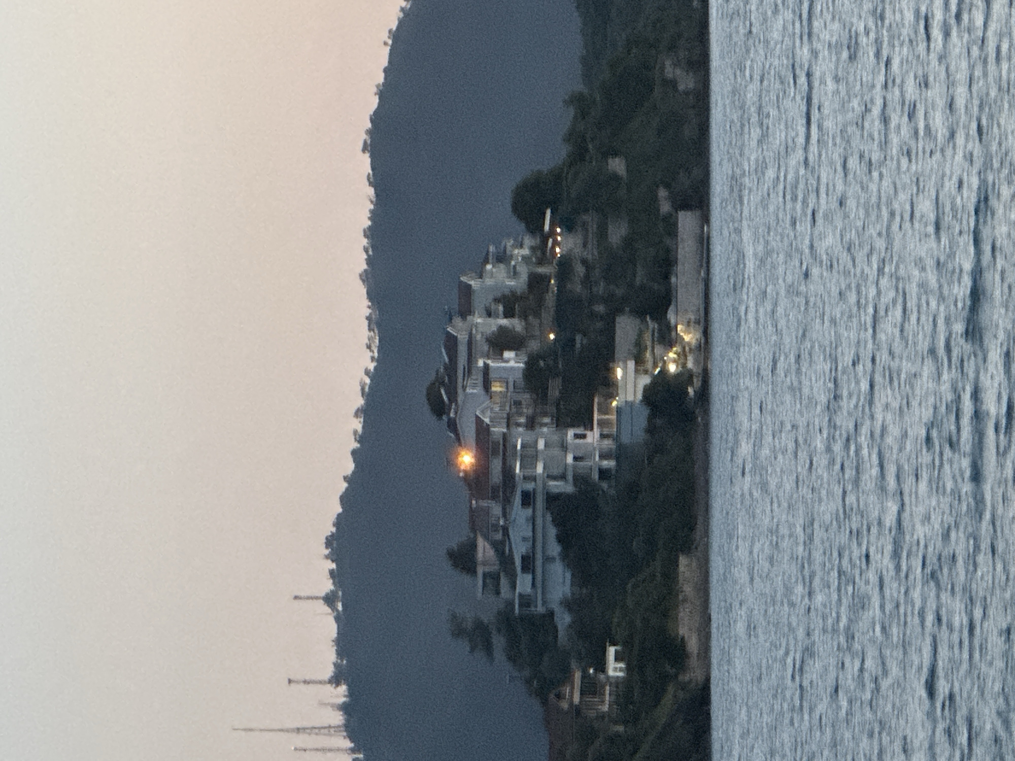 This media file captures a serene coastal scene at dusk or early night in Fethiye, Turkey. A large body of water, likely the sea or a bay, occupies the foreground, its surface showing texture from small waves or reflections. On the opposite shore, a densely built settlement of white and light-colored buildings with various illuminated windows and outdoor lights sprawls across a dark, tree-covered hillside. These structures appear to be residential properties or resort accommodations, many featuring modern architectural elements.
The hillside itself is rugged, covered in dark vegetation, and rises steeply from the water. In the background, a soft, muted light beige sky suggests twilight, with the last vestiges of daylight fading. Faint, tall, dark vertical structures, possibly antennas or distant masts, are visible against the sky on the far left. The overall atmosphere is peaceful and calm, with no specific human activity discernible. No text is visible within the image.