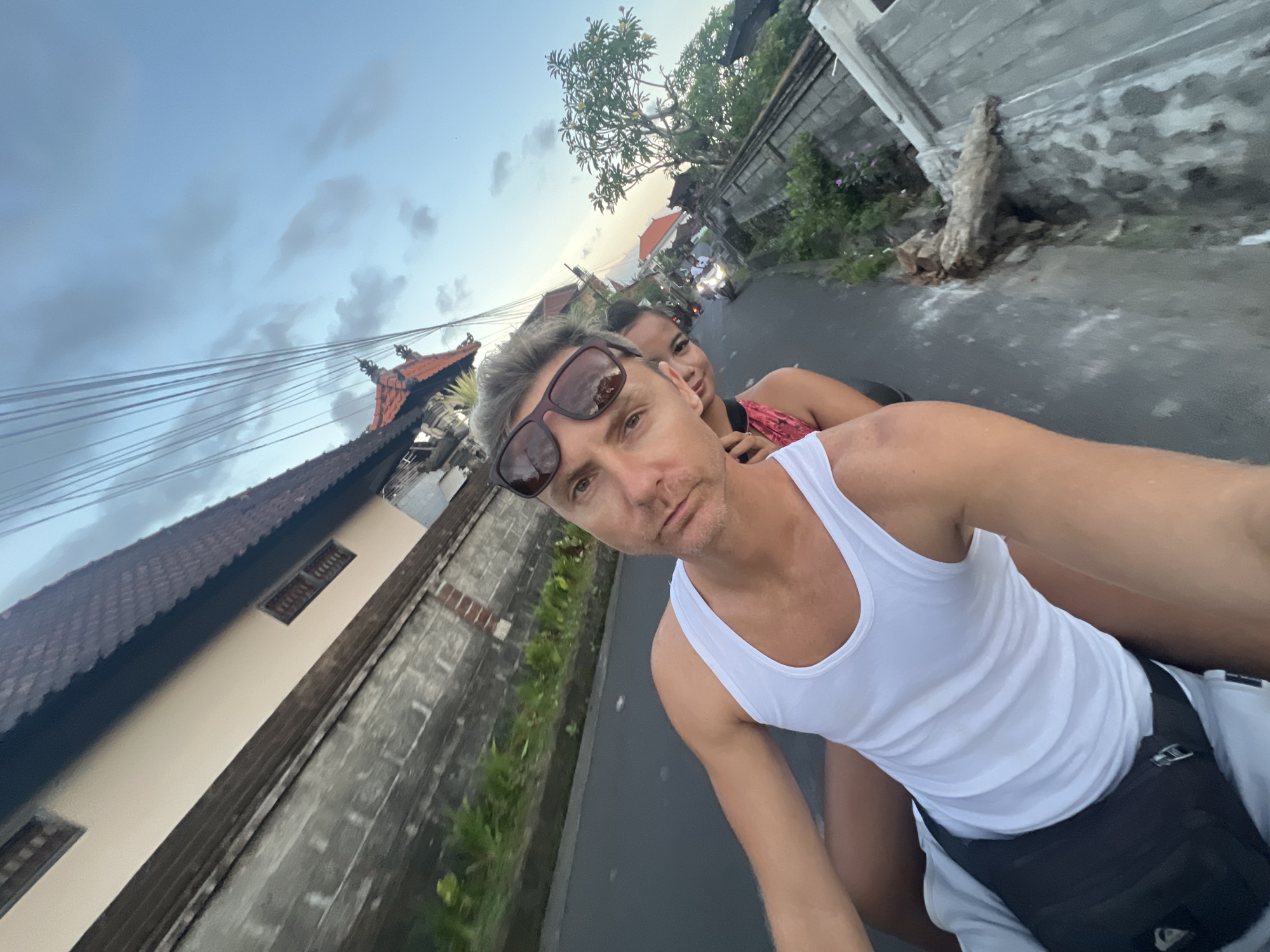 A selfie shows two individuals on a moving moped on a road in Sukawati, Indonesia. A fair-skinned man with grey hair and sunglasses wears a white tank top and black fanny pack. A darker-skinned woman with her hair up in a bun is riding behind him. They are on a narrow asphalt road flanked by high walls. The wall on the left is covered in green vegetation. Across the road is a grey cement wall with a tree root embedded in it. In the background, there are more mopeds on the road. The sky is cloudy. A building with a red tiled roof can be seen on the left, and greenery is visible above the walls on both sides.