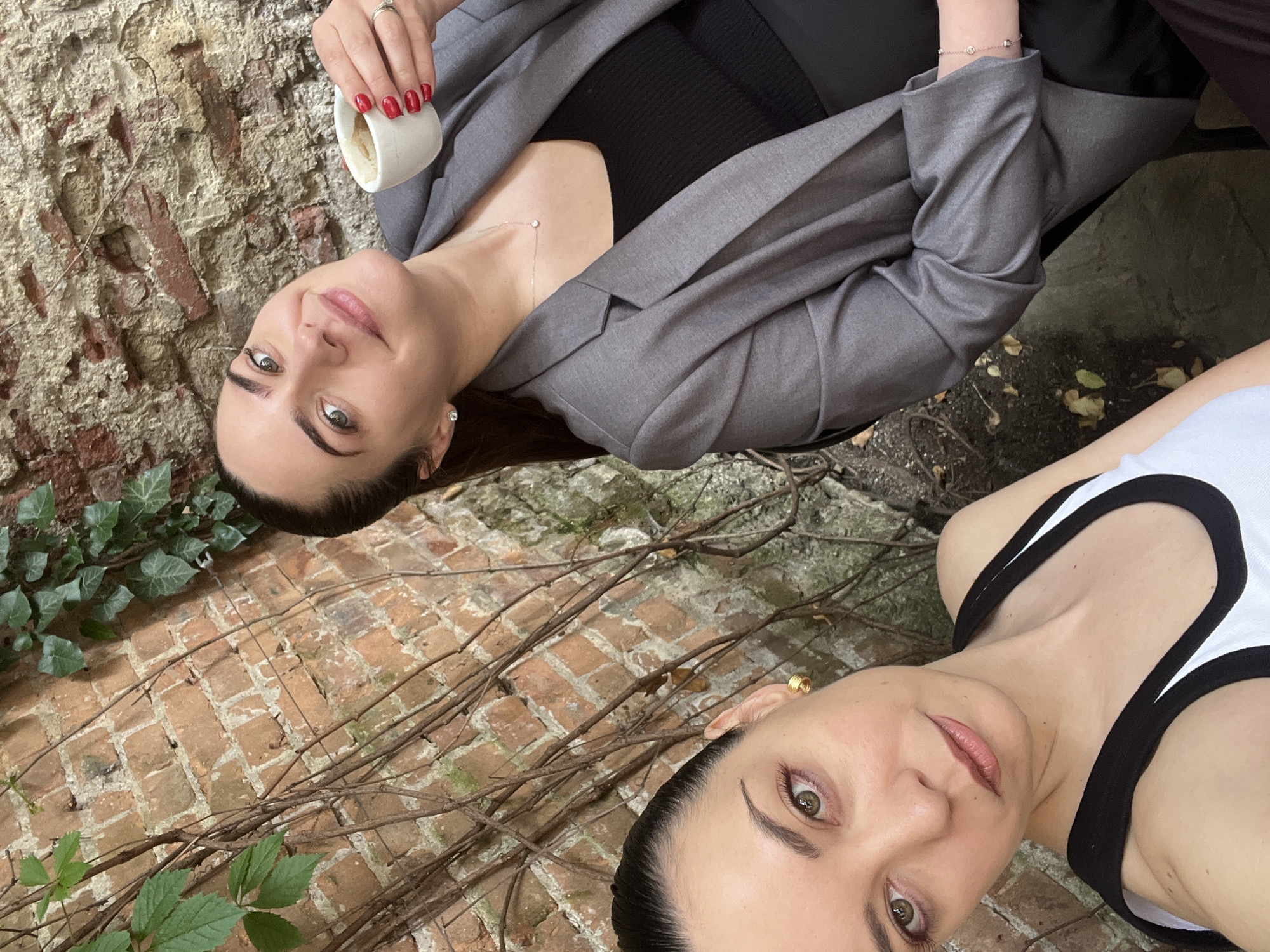 Two women are seated outdoors in front of a weathered brick wall partially covered with ivy. The woman on the right is holding a small white cup of coffee and smiling. She is wearing a gray blazer over a black top and a delicate necklace. The woman on the left is looking directly at the camera, and she is wearing a white tank top with black trim. The setting appears to be a courtyard or patio area, possibly in Sofia, Bulgaria, given the architectural style of the brickwork and the general ambiance.