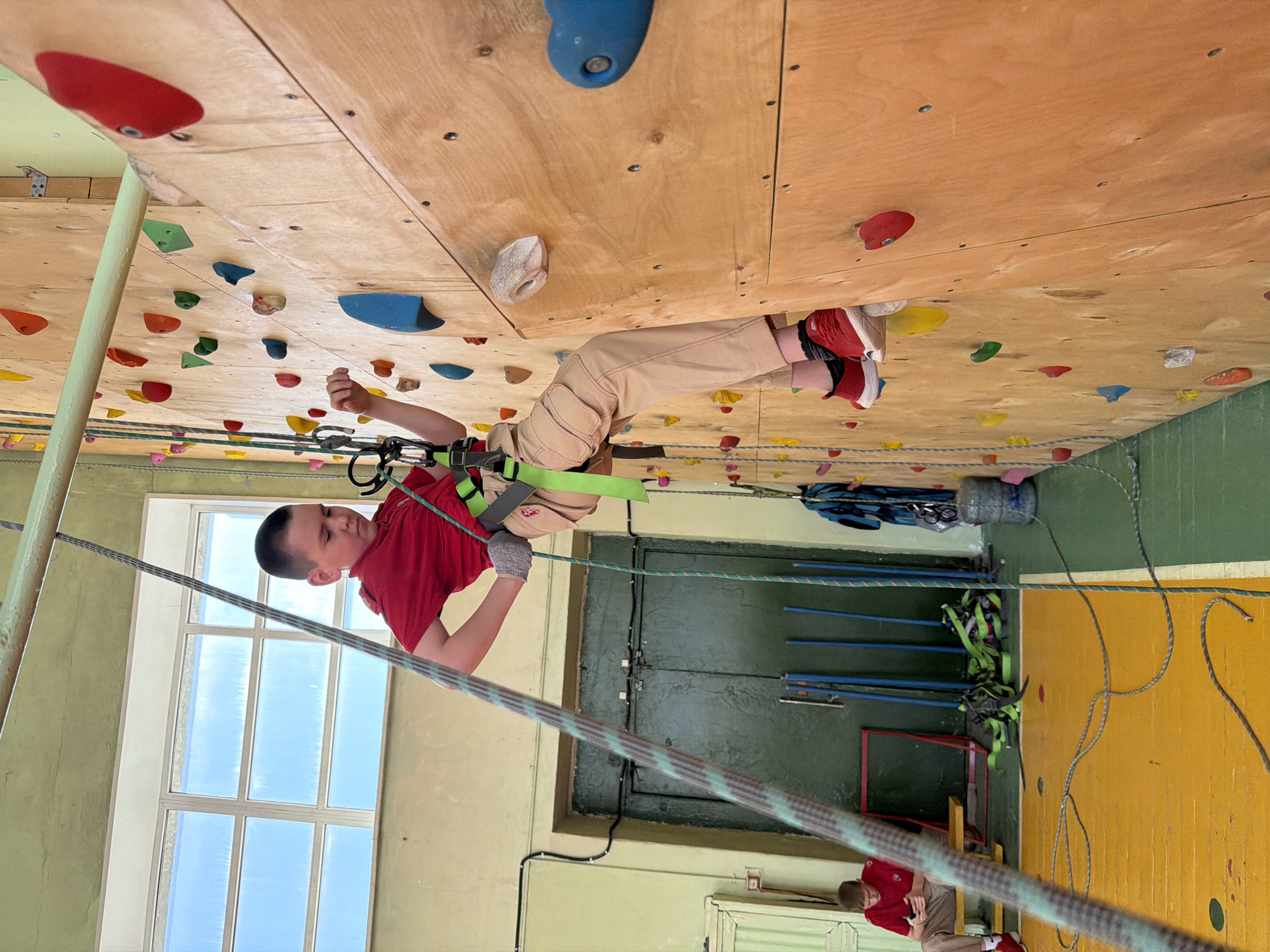 A child is suspended upside down on a climbing wall inside a room with a green and yellow painted floor. The child is wearing a red shirt, tan pants, and red shoes. A harness is secured around their waist, and they are holding onto a rope. The climbing wall is made of plywood and has various colorful climbing holds attached to it. A window is visible in the background. Another person is present, standing near a doorway, wearing a similar red shirt and tan pants. A collection of climbing gear, including carabiners, straps, and ropes, is visible on the floor near the climbing wall. The location of the image is Izhevsk, Russia.