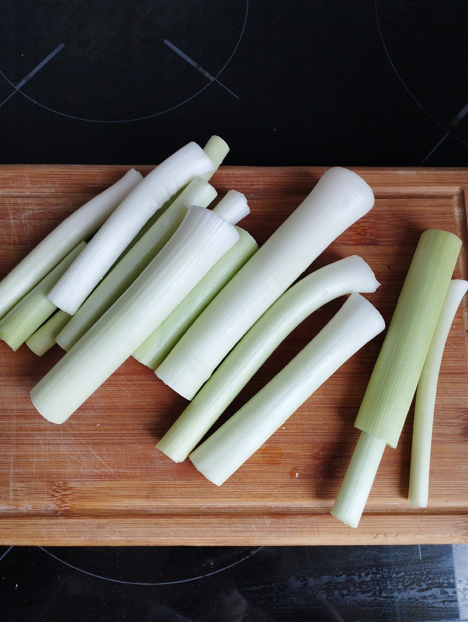 On a wooden cutting board, there are approximately a dozen pieces of cut leeks, varying in length and thickness. The leeks range in color from white to light green. The cutting board has a worn appearance with visible scratches and is placed on top of a black surface. The exact location in Burgas, Bulgaria, cannot be determined from the provided image.