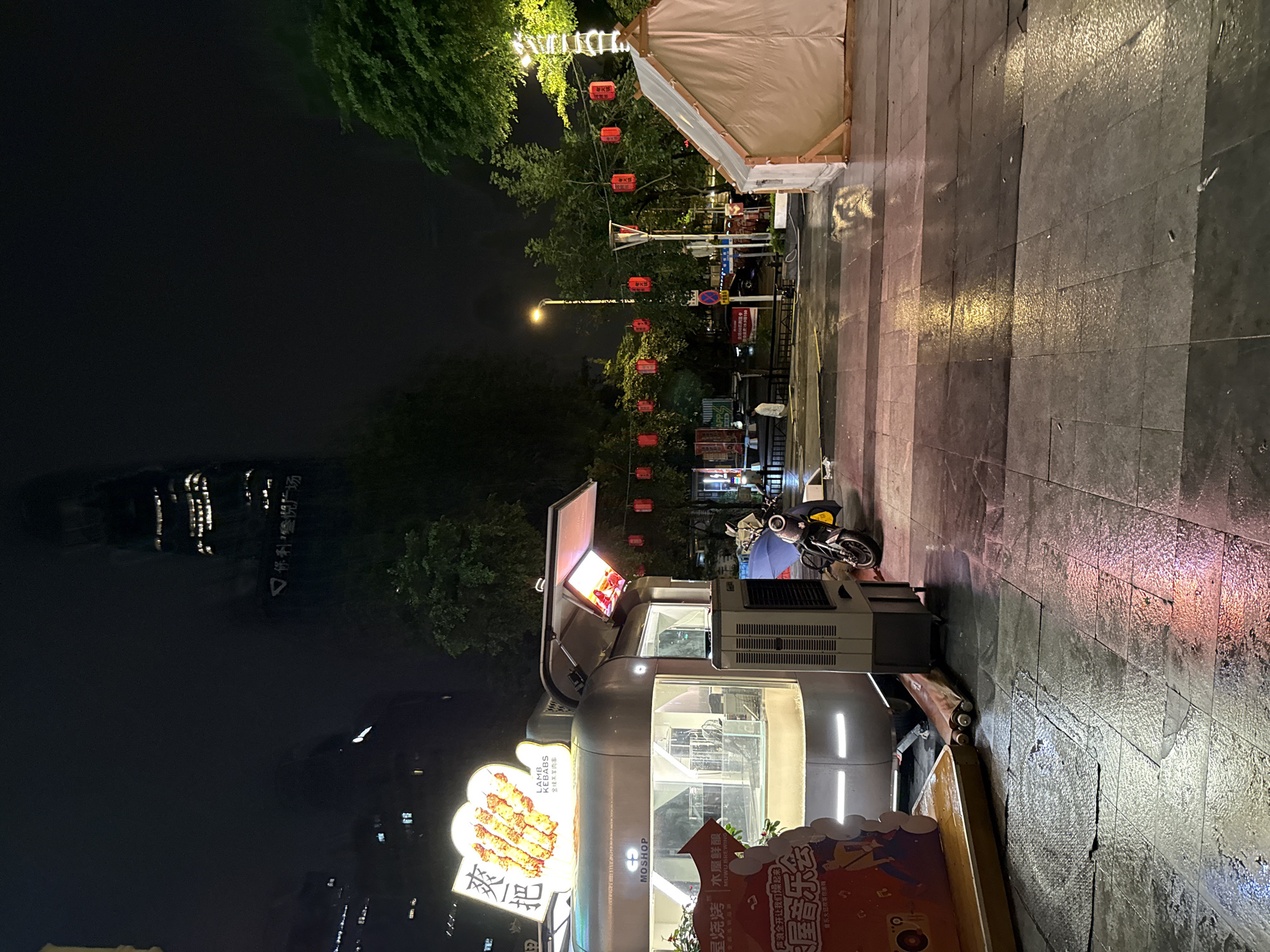 At night in Haizhu District, China, a food stall with a sign displaying "LAMB KEBABS" sits on a wet paved street. A motorcycle is parked next to the stall, and a blue umbrella is propped up. In the background, red lanterns are strung across trees, and a white tent is set up. A tall building with illuminated windows is visible in the distance, partially obscured by fog. The streetlights and the stall's lights cast reflections on the wet pavement.