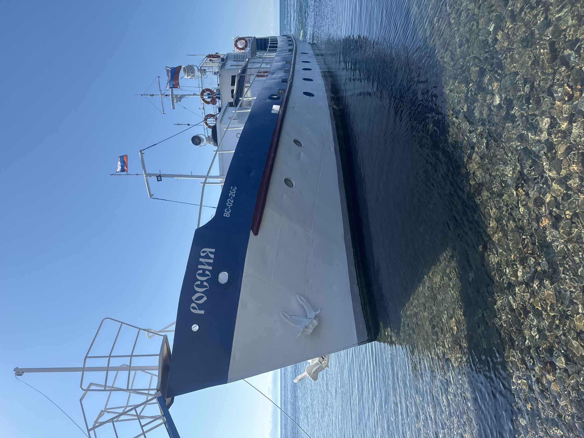 A white and blue passenger boat, named "РОССИЯ" (Russia), is moored in shallow, clear water on a sunny day. The boat is positioned at an angle, with its bow facing towards the viewer and slightly to the left. The water is so clear that the rocky seabed is visible beneath the surface. Two Russian flags are flying on the boat's masts. The boat appears to be docked near the shore of a lake or large body of water, with a shoreline visible in the distance. The sky is a clear, bright blue, indicating fair weather. The scene suggests a calm and serene environment, likely a tourist destination. The location is identified as Listvyanka, Russia, a settlement on the western shore of Lake Baikal.