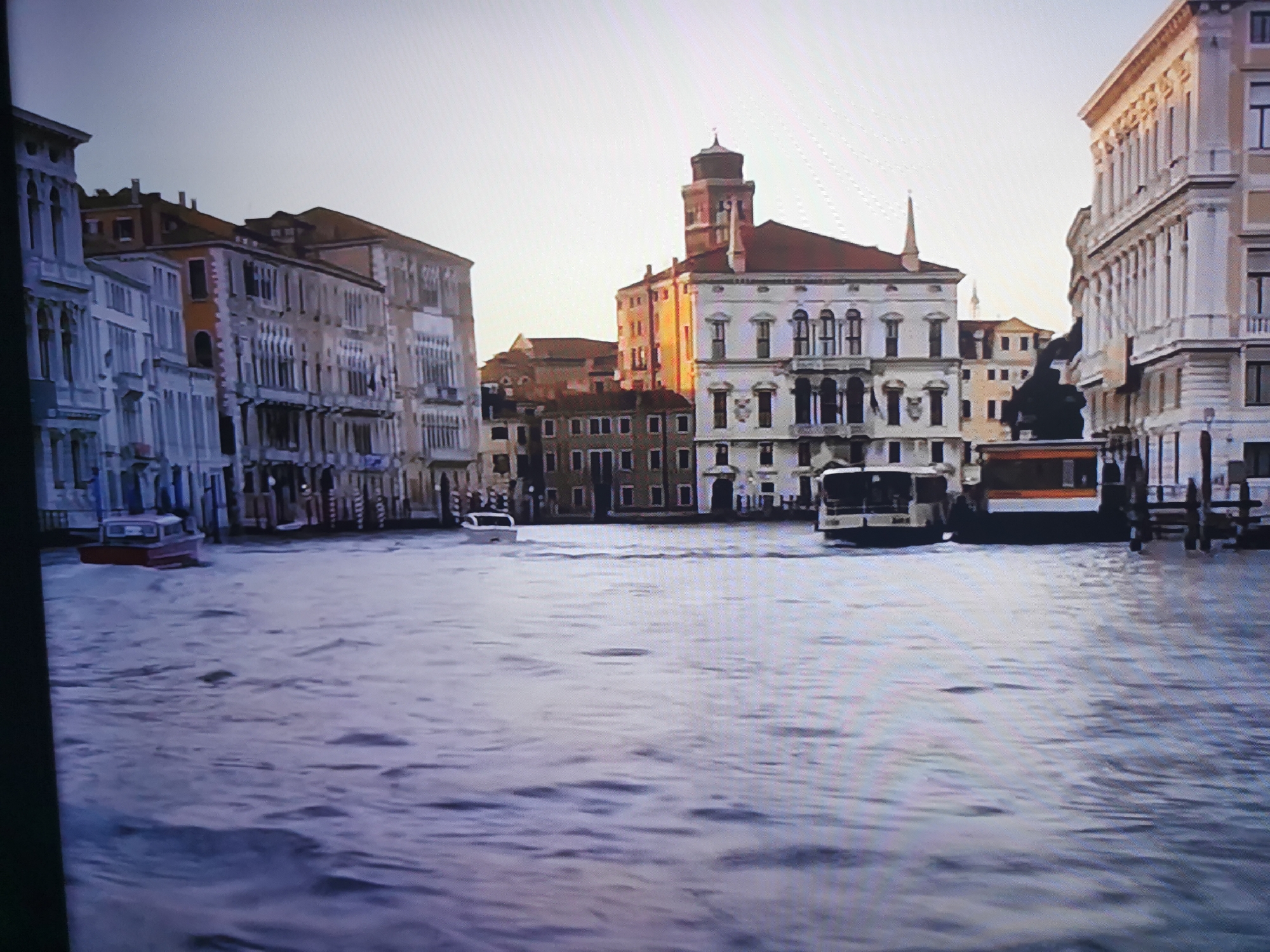 The image depicts a flooded canal with several boats navigating the water. On either side of the canal are tall, multi-story buildings with ornate architecture. The sky is a pale, overcast gray. One boat, colored red and white, is in the foreground, appearing to be a small motorboat. Further in the distance, larger vessels, possibly tour boats or ferries, are visible. The water level is high, reaching the lower floors of some buildings. The scene appears to be a historic European city, likely Venice, Italy, known for its canals and flooding. The city of Dnipro, Ukraine, is not depicted in this image.