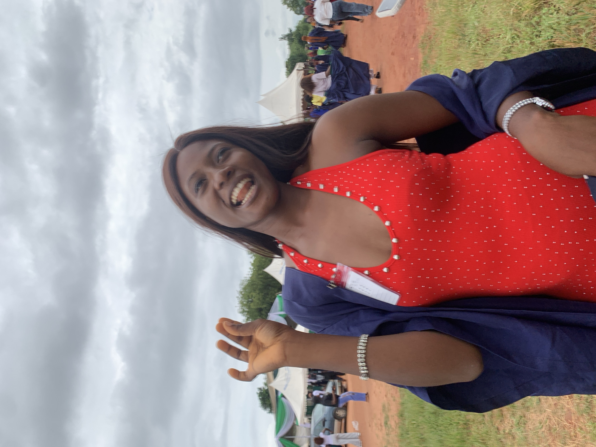 The image shows a smiling woman outdoors, likely in Ukwu-Abwa, Nigeria, given the location request. She is wearing a red top with white embellishments and a navy blue garment draped over her shoulders. A silver bracelet is visible on her wrist. Her hand is raised, perhaps in a wave or gesture. A nametag is attached to her clothing. In the background, there is a gathering of people, some wearing similar navy blue garments. Tents and vehicles are visible, indicating an event. The ground appears to be a mix of grass and red dirt. The sky is overcast.