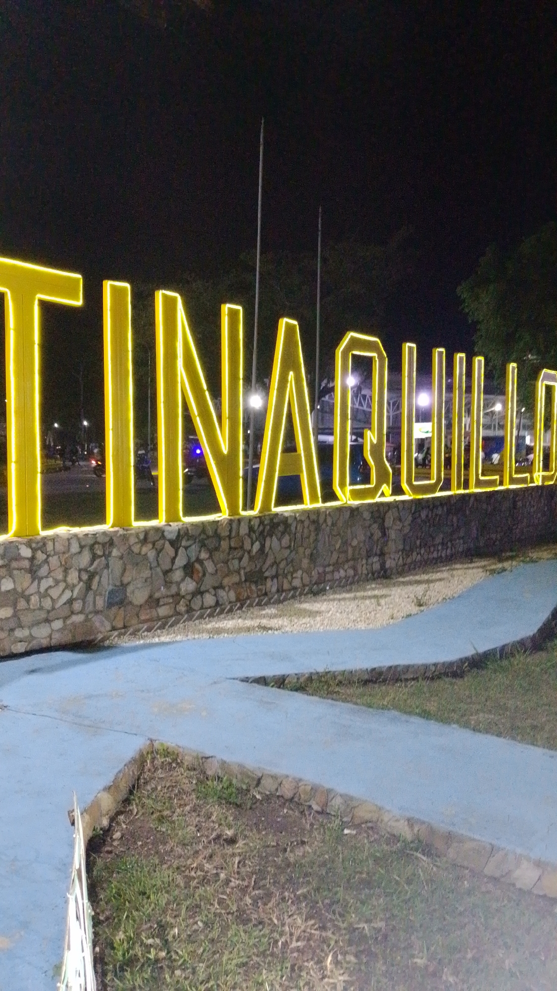 At night, a brightly lit sign spells out "TINAQUILLO" in large yellow letters. The letters are mounted on a low wall made of stone. A pale blue pathway leads up to the sign. Beyond the sign, there are trees, streetlights, and some blurry lights from vehicles. The ground in the foreground is covered with grass and some bare patches of earth. The location is likely in Tinaquillo, Venezuela, based on the sign.