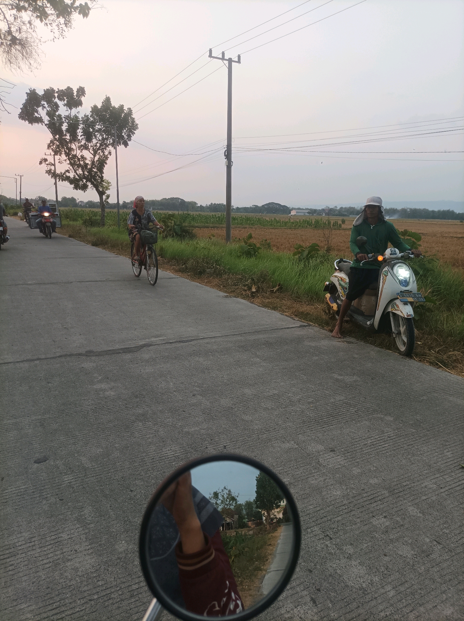 The image depicts a rural road in Canga'an (DK. Jetis), Indonesia, during what appears to be either sunrise or sunset, given the soft, diffused light. The road, made of concrete with visible expansion joints, curves gently to the left. On the right side of the road, a person wearing a green long-sleeved shirt and a white hat is seated on a white scooter parked on the grassy shoulder, facing forward. Their left leg is extended, and their right leg is bent. Behind them, a harvested field of dry straw stretches to the horizon, with a line of trees and vegetation bordering it.
On the left side of the road, a woman is riding a bicycle towards the viewer. She is wearing a patterned dress and a headscarf. Further up the road, two motorcycles are visible, one with a sidecar carrying cargo and a rider. The environment is characterized by scattered trees, power poles with overhead wires, and the distant outline of hills or mountains. In the foreground, the reflection in a circular mirror, likely from the viewer's own vehicle, shows a portion of an arm and shoulder wearing a reddish-brown garment. The sky is a pale, hazy color, suggesting overcast conditions or atmospheric haze.