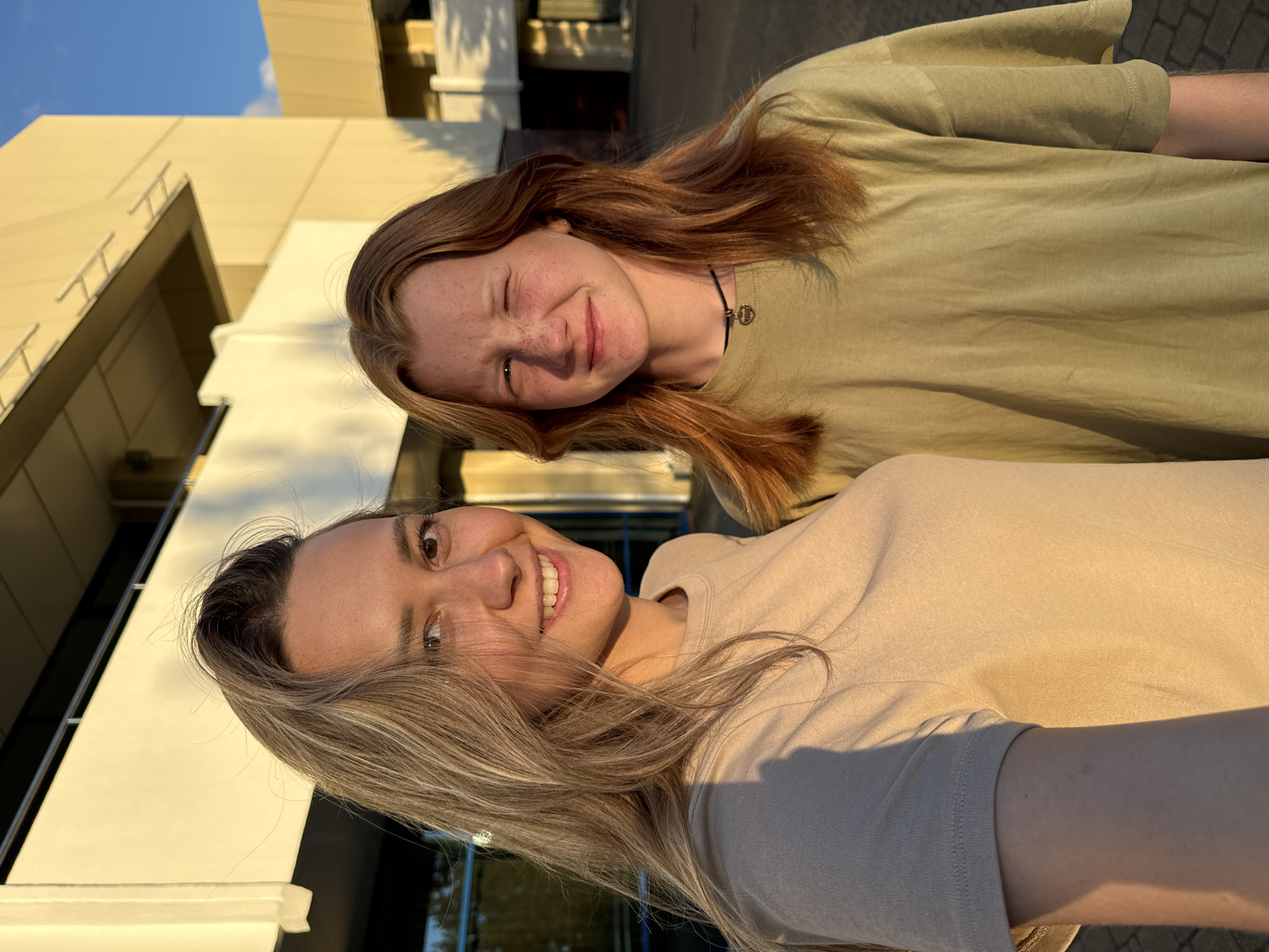 This is a close-up, rotated selfie of two young women outdoors. The woman in the foreground has fair skin, prominent freckles, and is smiling broadly with her eyes open. She has light brown hair with blonde highlights, and is wearing a light beige t-shirt. The woman behind her, slightly out of focus, has red hair and is winking, also with a smile. She has freckles as well and is wearing a muted green t-shirt. They appear to be in a casual outdoor setting, possibly near a modern building with cream-colored walls and windows, suggested by the background. The lighting indicates it is late afternoon or early evening, with the sun casting a warm glow. The environment seems relaxed and friendly.
