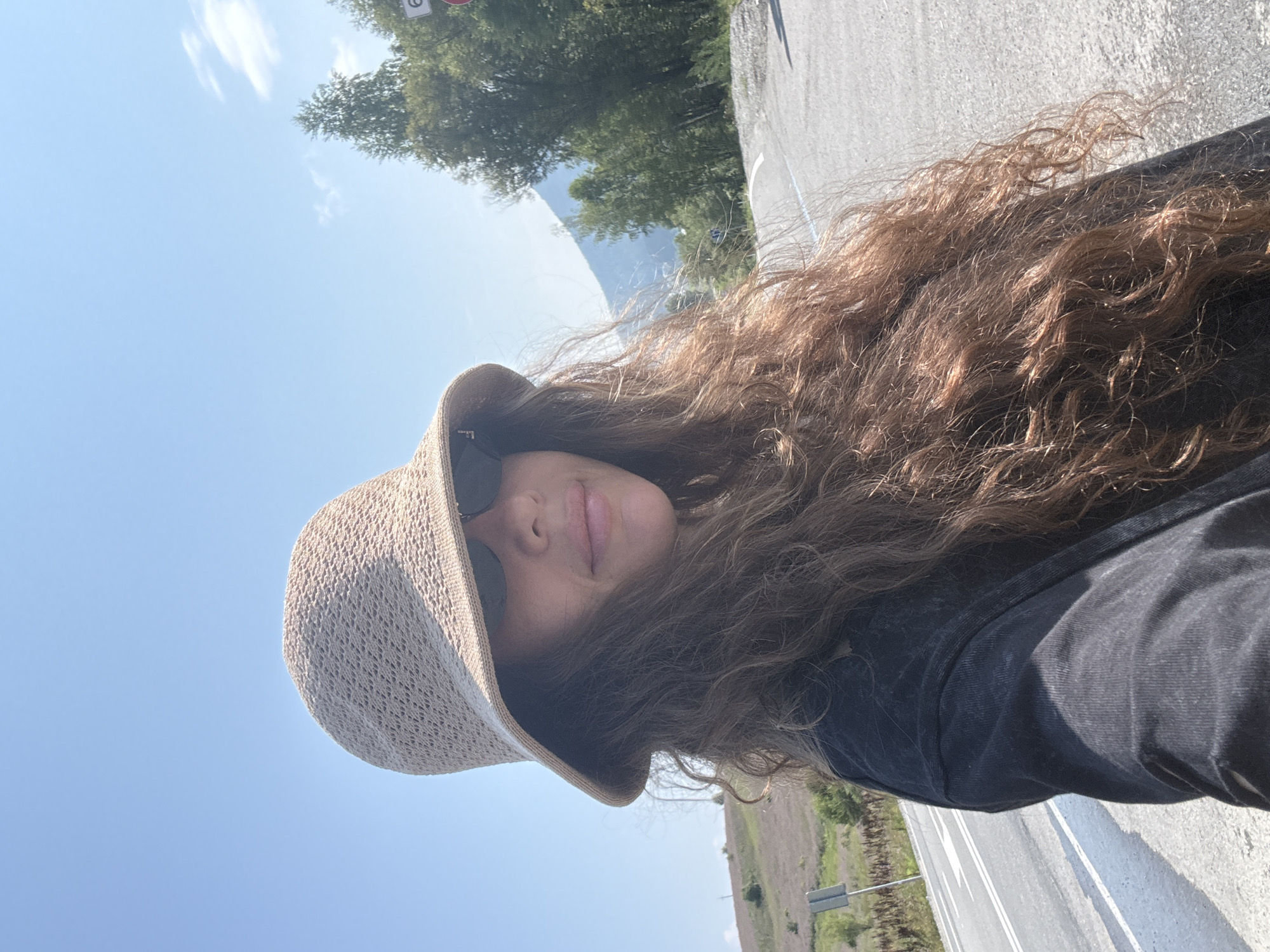 A woman wearing a wide-brimmed straw hat and sunglasses poses for a selfie outdoors. She has long, curly brown hair and is wearing a dark t-shirt. The background shows a paved road with white markings, a grassy hillside, and trees. The sky is clear and blue. The image appears to have been taken in a rural or mountainous area, likely outside of a city. There are no visible landmarks or signs that would specifically place the location in the city of Choya, Russia.