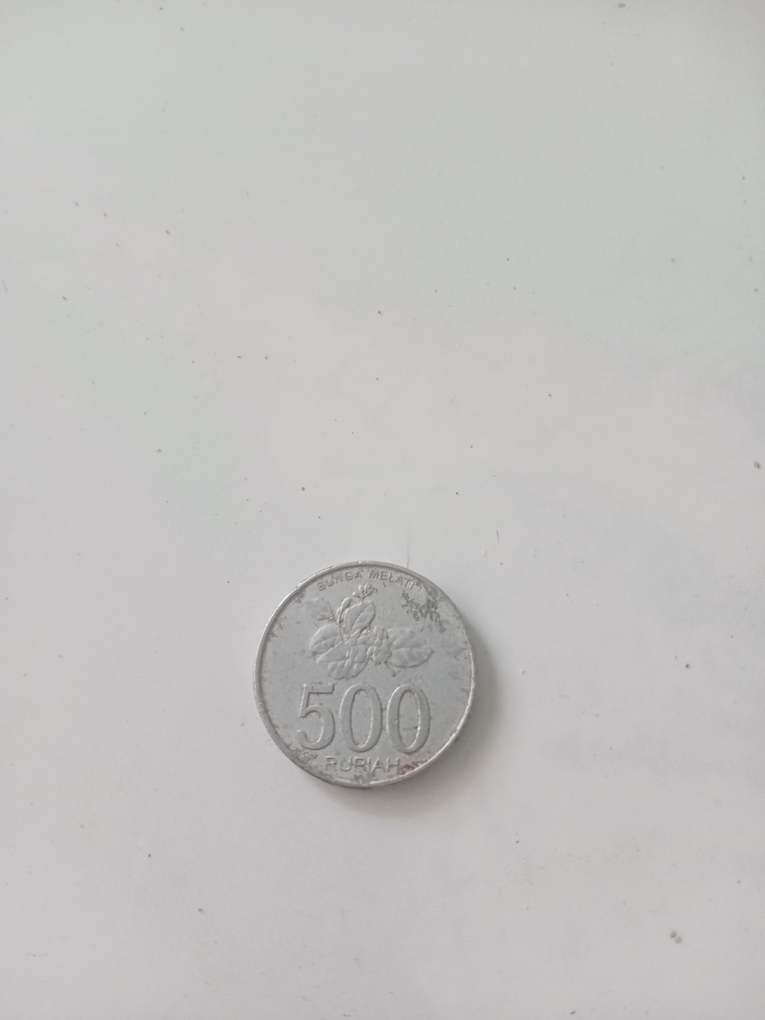 A silver coin with the numeral "500" embossed on it is pictured on a plain white surface. The coin also features a floral design and the inscription "BUNGA MELATI" and "RUPIAH". The scene is a close-up shot, and no specific location in Badas, Indonesia, is depicted.