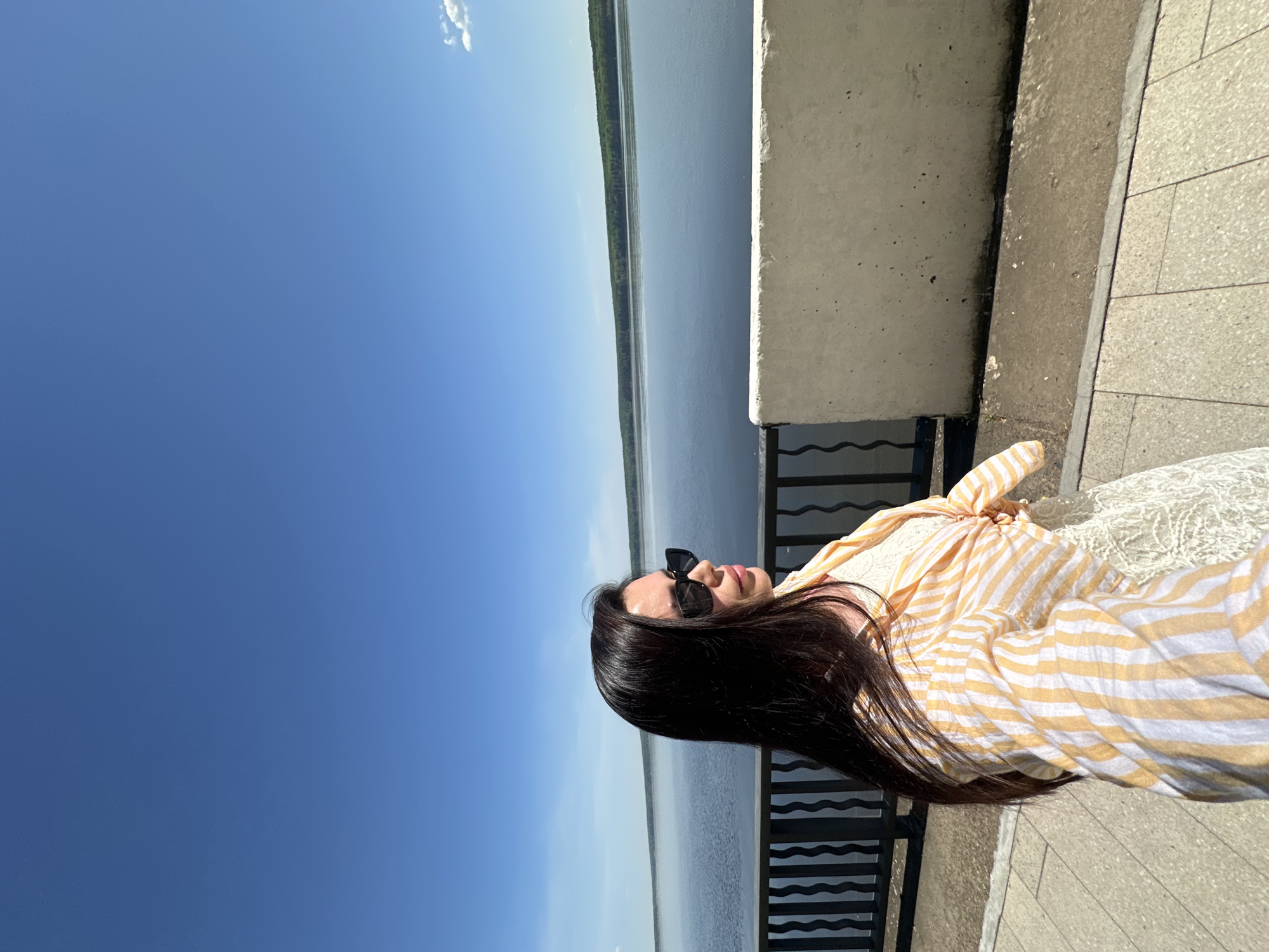 A woman is standing on an elevated walkway adjacent to the river Kama in Chaikovsky, Russia. The river extends into the distance, bordered by a line of trees on the opposite bank. The sky is clear and blue. The woman has long, dark hair and is wearing sunglasses and a striped yellow and white shirt. A section of a concrete wall and a metal railing separate the walkway from the river. The walkway is paved with light-colored stones.