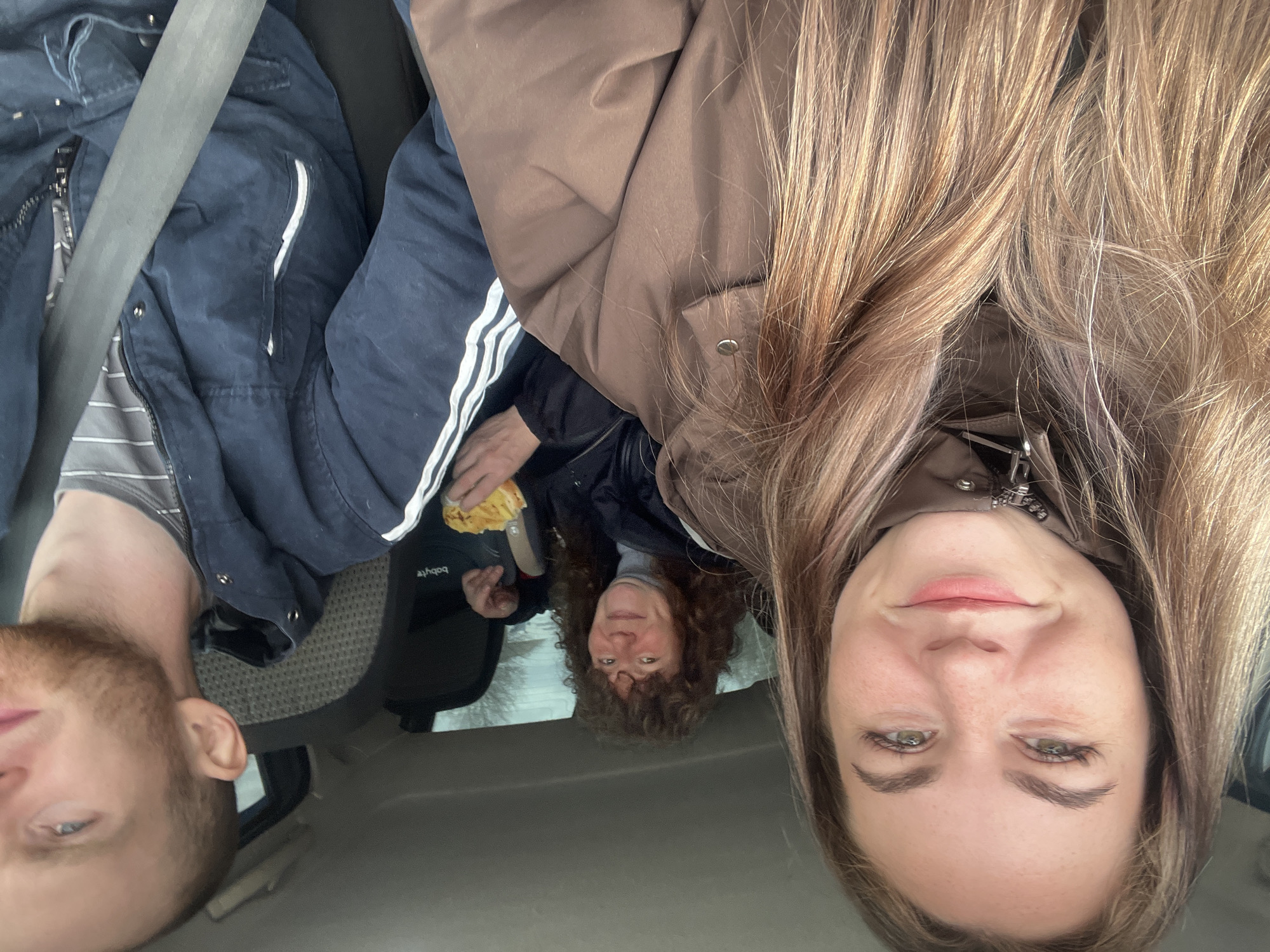 Three people are in a vehicle, likely a car, as indicated by the interior ceiling. The person on the left has short brown hair and a beard, wearing a striped shirt and a blue jacket with white stripes. The person on the right has long blonde hair and is wearing a brown jacket. In the back, a woman with curly brown hair sits in a car seat, holding something yellow. The location of Chaikovsky, Russia, is not visually confirmable from the provided image.