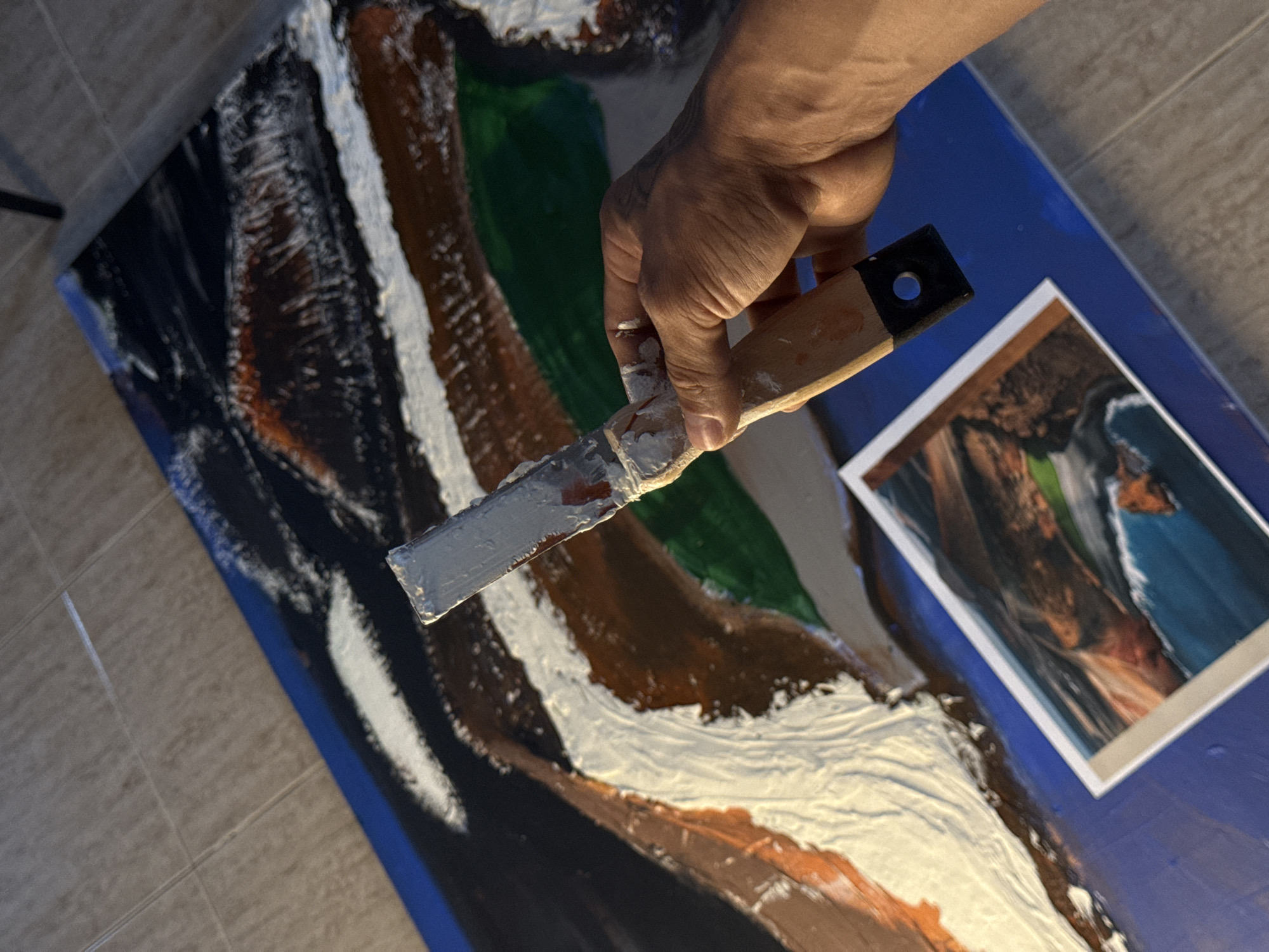 A person's hand holds a paint scraper over an abstract painting on a blue surface. The painting contains black, white, brown, and green paint in thick layers. A photographic print rests on the blue surface next to the painting. The photo and painting depict a similar scene, possibly a mountain range or landscape with similar colors. The scene is located indoors, as indicated by the floor tiles. There is no information on the location in the city of Yaiza, Spain.