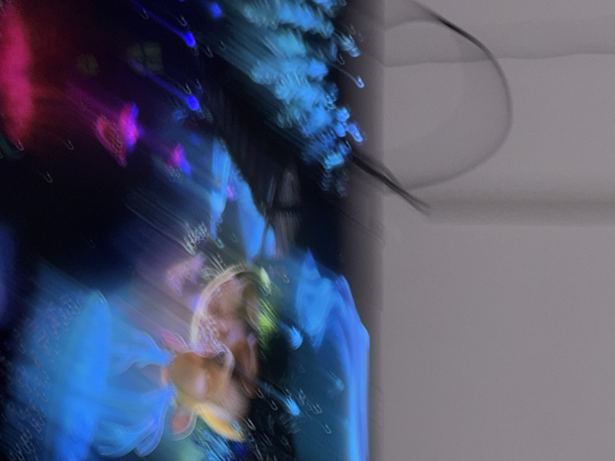 The image depicts a blurred, abstract scene with vibrant neon lights. In the foreground, a deer-like creature with glowing antlers is visible, lying down and appearing to be asleep. The background is filled with streaks of blue, pink, and purple light, suggesting a dynamic and possibly nocturnal environment. There are no discernible people or specific architectural features that would allow for pinpointing a location within Skopje, North Macedonia. The overall impression is one of fantasy or a stylized representation of a magical forest.