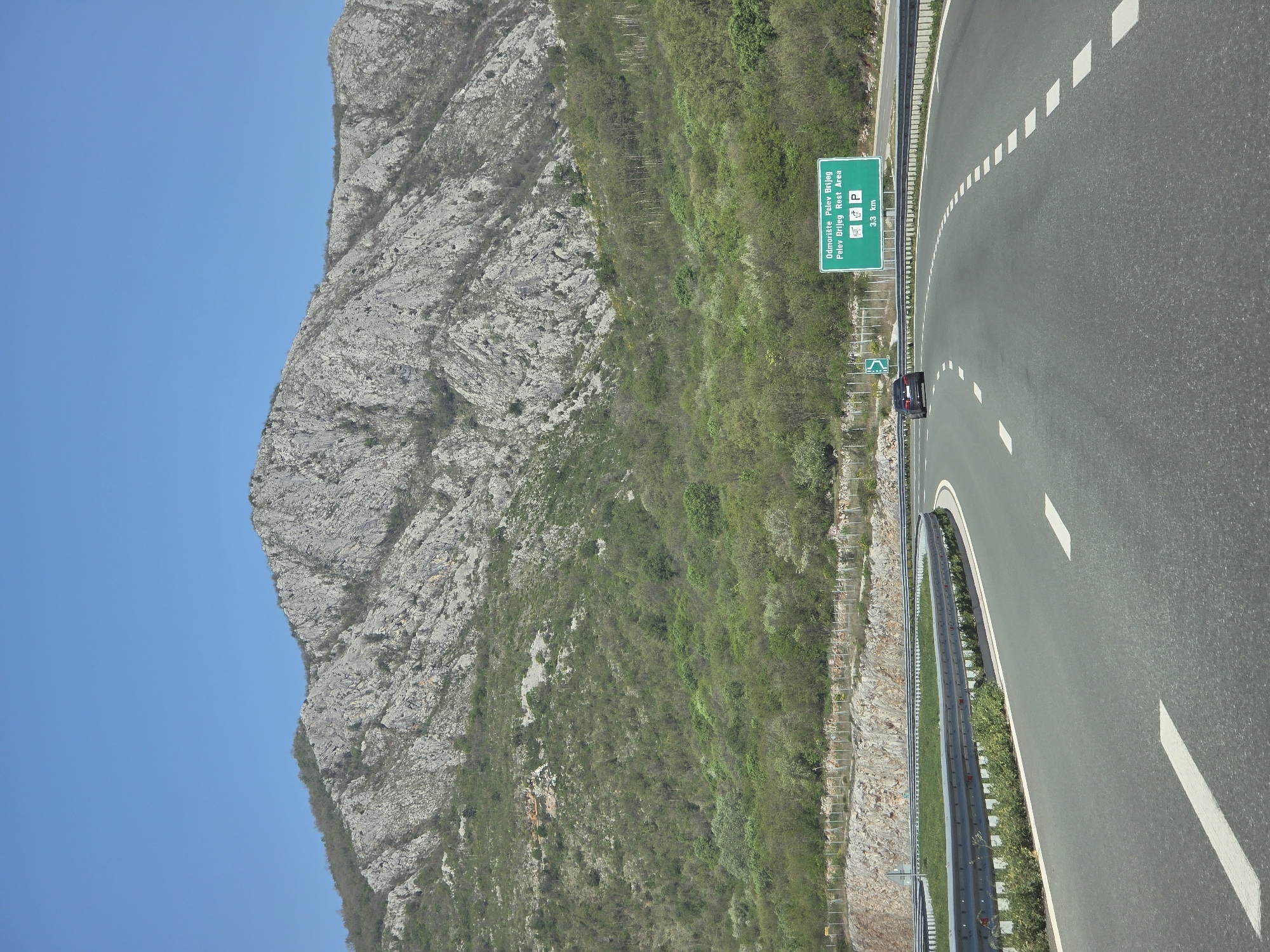 The image depicts a highway in Pelev Brijeg, Montenegro, adjacent to a mountain. The road curves slightly, with white lane markings. A sign indicates a rest area "Odmorište Pelev Brijeg" with parking and restroom facilities. A single car is visible on the road. A crash barrier separates the road from the mountainous terrain. The mountain is rocky and covered in sparse vegetation. The sky is clear and blue.