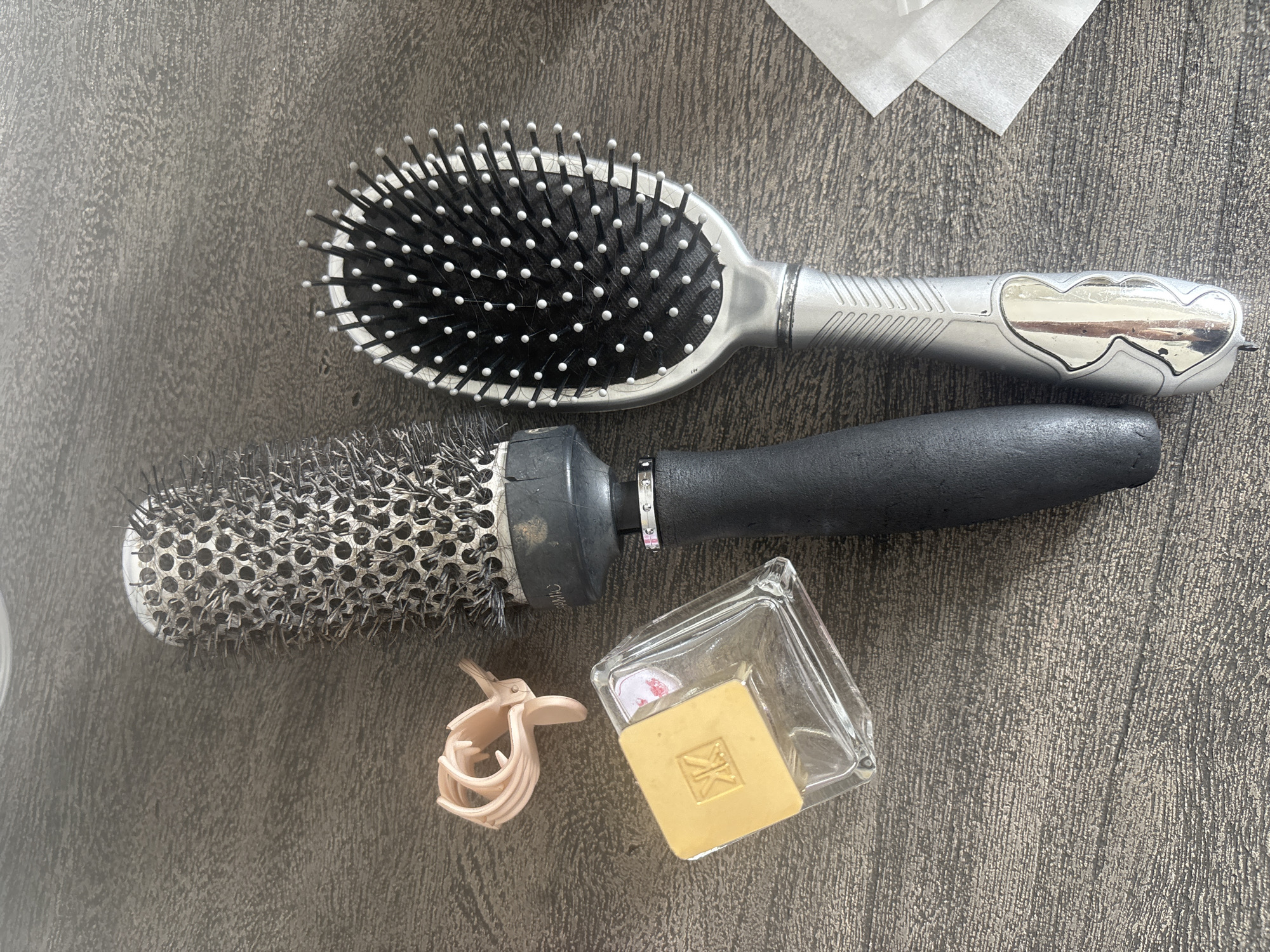 This image depicts a still life composition of hair styling tools and a small glass perfume bottle, arranged on a textured, dark gray surface, possibly a wooden table. From left to right, a small, light pink plastic hair clip is partially visible. Adjacent to it is a clear glass perfume bottle with a square, gold-colored cap featuring embossed markings. Next to the bottle is a large, black-handled round brush with silver bristles and a ceramic barrel, angled slightly. To the right of this brush is a silver-handled oval paddle brush with black bristles tipped with white. A small stack of white paper tissues or wipes is located in the lower right corner. The lighting suggests an indoor setting, and the overall scene is static, with no individuals or animals present. The image does not provide any information to determine its location in Dubai, United Arab Emirates.