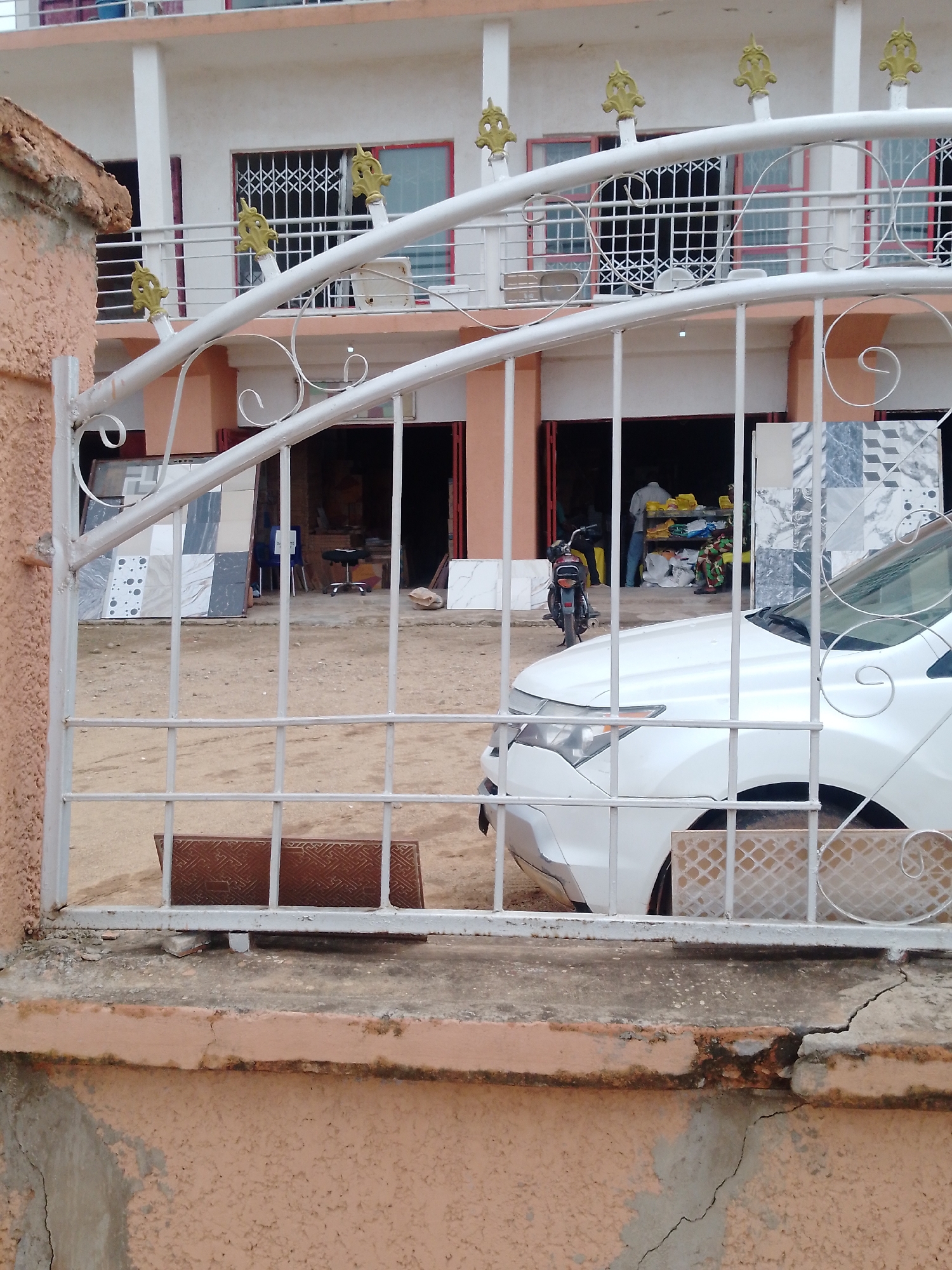 The image, taken in Sabon Tasha, Nigeria, shows a view through a white metal gate into an open-fronted business. Inside the business are stacks of tiles, a motorcycle, a car parked partially blocking the view, and several people. The building behind the business is two stories tall with white walls and a reddish-brown trim around the window and door openings. The gate is mounted on a low wall, which is showing signs of cracking.