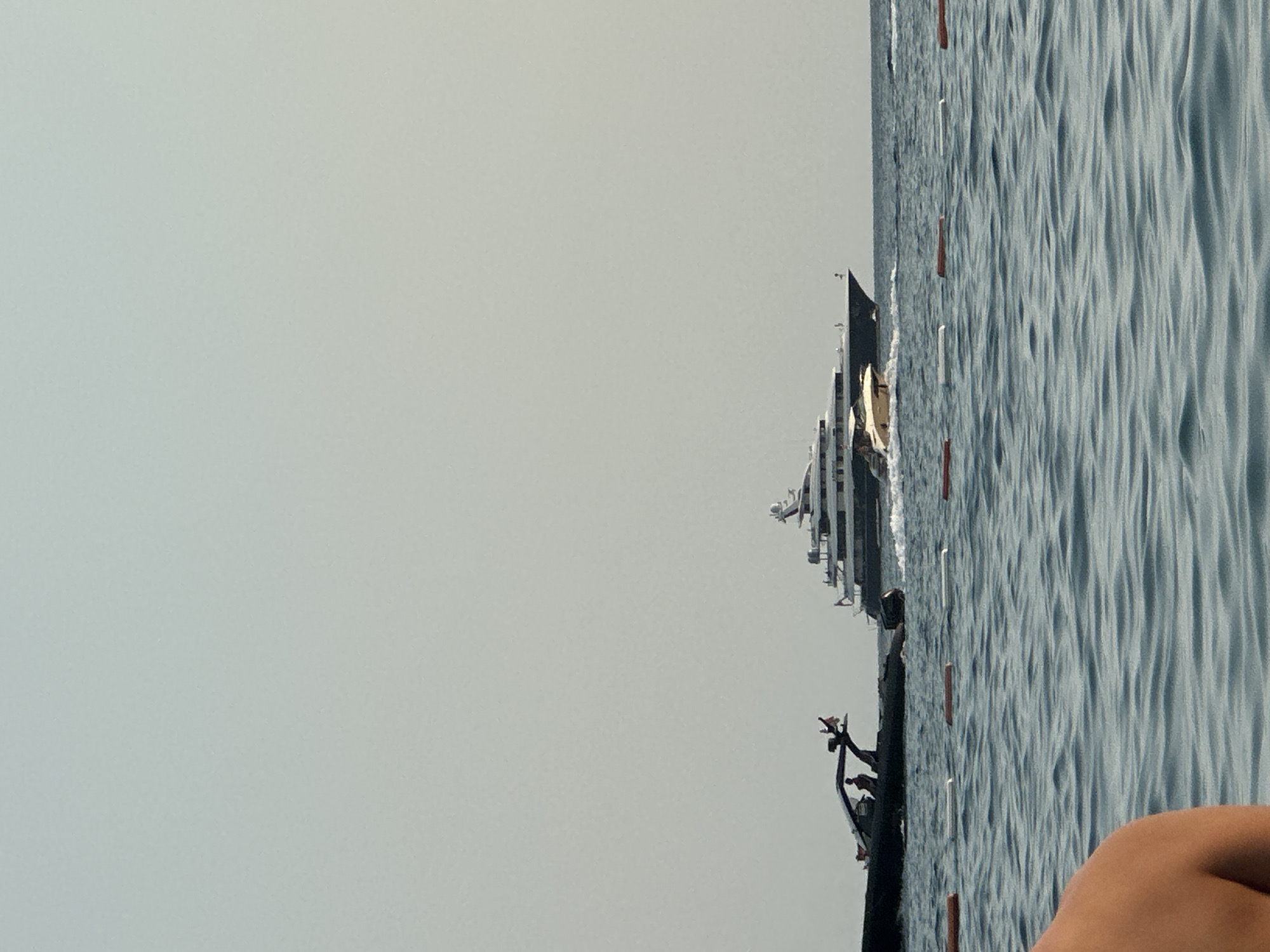 A wide shot captures a vast expanse of calm, blue water in the foreground, with gentle ripples disturbing its surface. In the mid-ground, a large, dark-hulled yacht with multiple decks and white superstructure is anchored in the distance. To the left of the large yacht, a smaller, white motorboat is in motion, creating a small wake. Further to the left, a sleek, black rigid inflatable boat (RIB) is also moving, with two individuals visible on board. A second, smaller white boat is also visible in the mid-ground between the RIB and the large yacht. Orange and white buoys are spaced out in the water, marking a boundary. In the extreme foreground, the shoulder and upper back of a person are visible, cropped from the left side of the frame. The sky above is a uniform, pale grey, suggesting an overcast or hazy day. The scene is set on the open water. This image appears to be taken from a beach or a boat near the coast of Boreti, Montenegro.