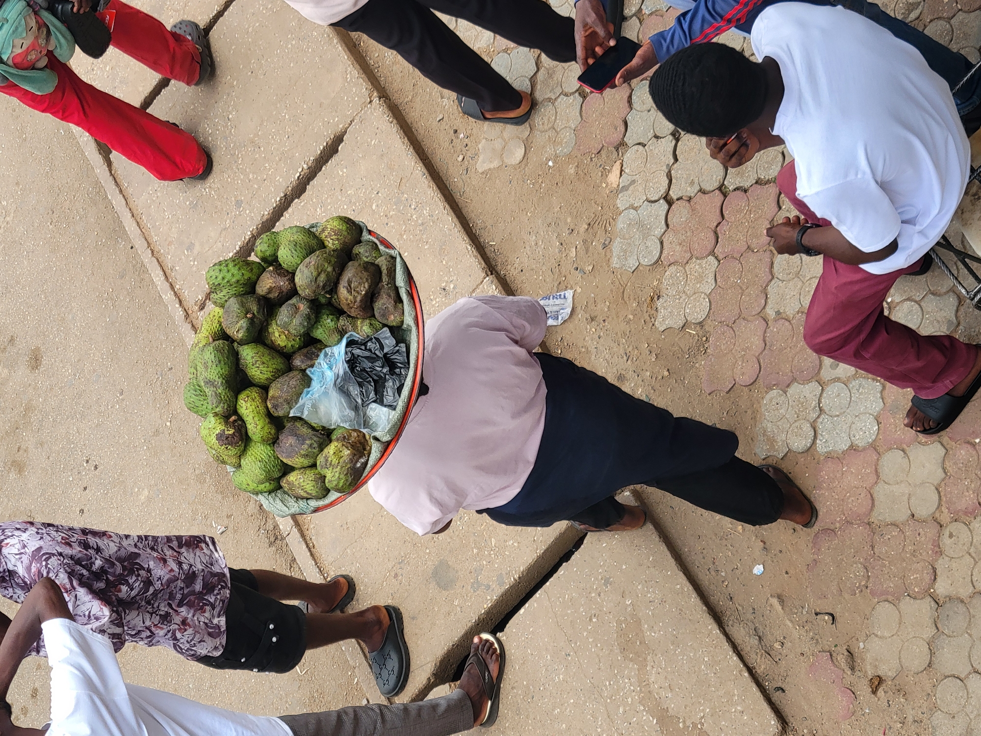 An overhead shot shows a street scene in Jos, Nigeria, with at least five individuals surrounding a woman who is selling a tray of soursop fruit. The woman is wearing a light pink top and dark blue skirt with black leggings. The tray she carries on her head is full of green soursops, with a plastic bag partially covering them. Two men are standing close to her. One man wears a white shirt with a purple and black patterned shirt visible around his waist and black shorts. The other man wears a white t-shirt, red pants, and black sandals. Another man appears to be holding a phone. The ground is a mix of concrete and brick paving.