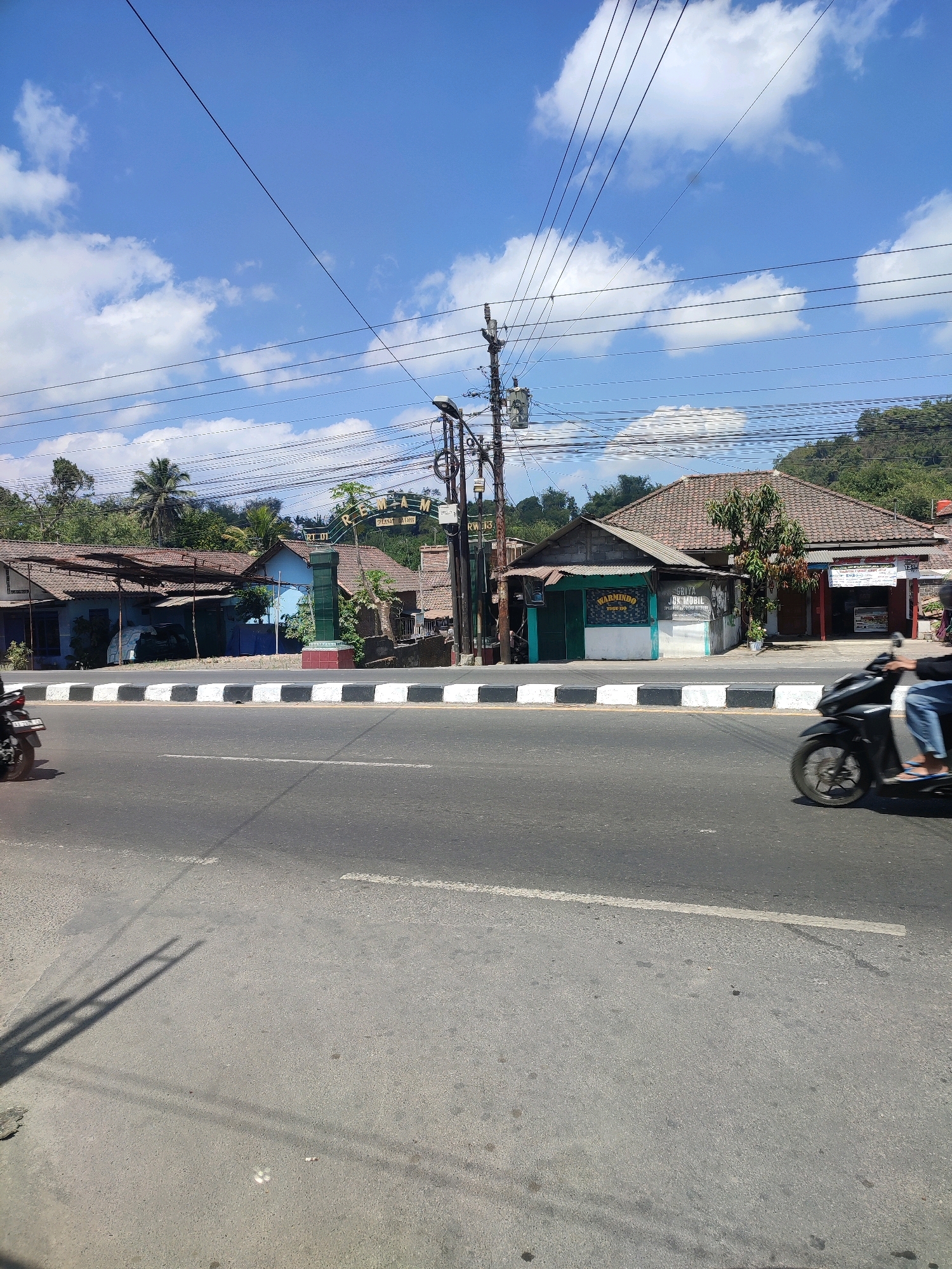 The image shows a street scene in Jumoyo, Indonesia. A paved road runs horizontally across the foreground, with a motorcycle in motion on the right side. A white and black striped curb separates the road from the sidewalk. Behind the curb, there are several buildings with tiled roofs, appearing to be shops or residences. An archway with text, possibly a gateway to a neighborhood, stands near the center of the image. Telephone poles and wires are visible against a bright blue sky with scattered white clouds. Trees and greenery are present in the background, and a hillside is visible in the far distance. The overall impression is of a typical day in a semi-urban or rural area.