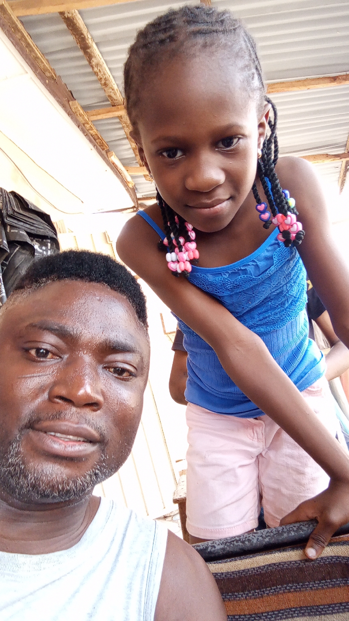 The image shows a man and a girl in Sabon Tasha, Nigeria. The man, likely in his 30s or 40s, has dark skin, short black hair, and a beard. He is wearing a white tank top. The girl, possibly around 8 years old, has dark skin and braided hair decorated with pink and white beads. She is wearing a blue tank top and pink shorts. They appear to be indoors, under a corrugated metal roof. The man is likely taking a selfie with the girl. A stack of black bags is visible in the background.