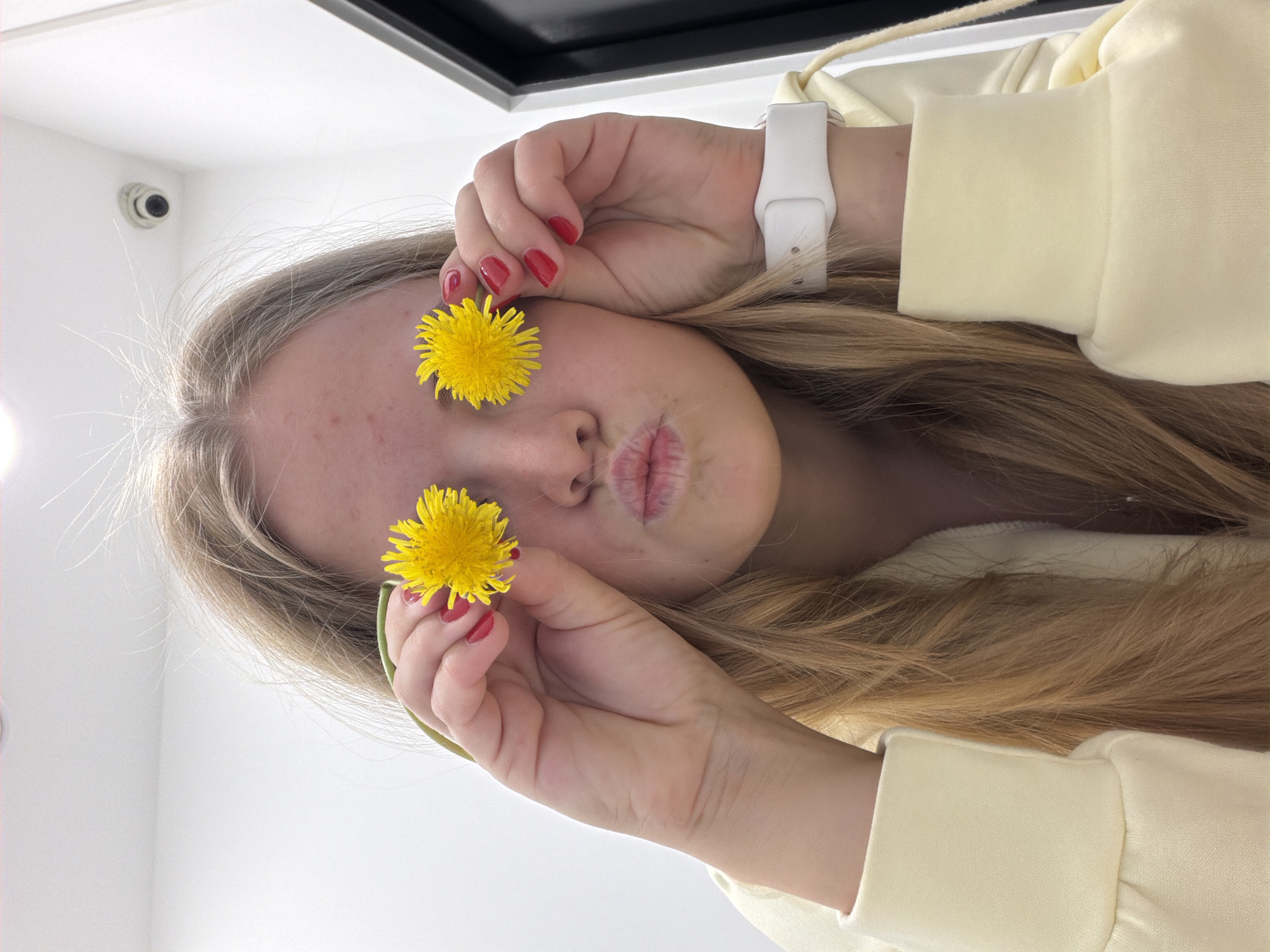 A young woman with long blonde hair is holding two yellow dandelions in front of her eyes. She is wearing a pale yellow sweater and a white smartwatch. Her nails are painted red. The background is a white ceiling with a black rectangular object and a white security camera in the upper left corner. It is not possible to confirm the location in Izhevsk, Russia from the given image.