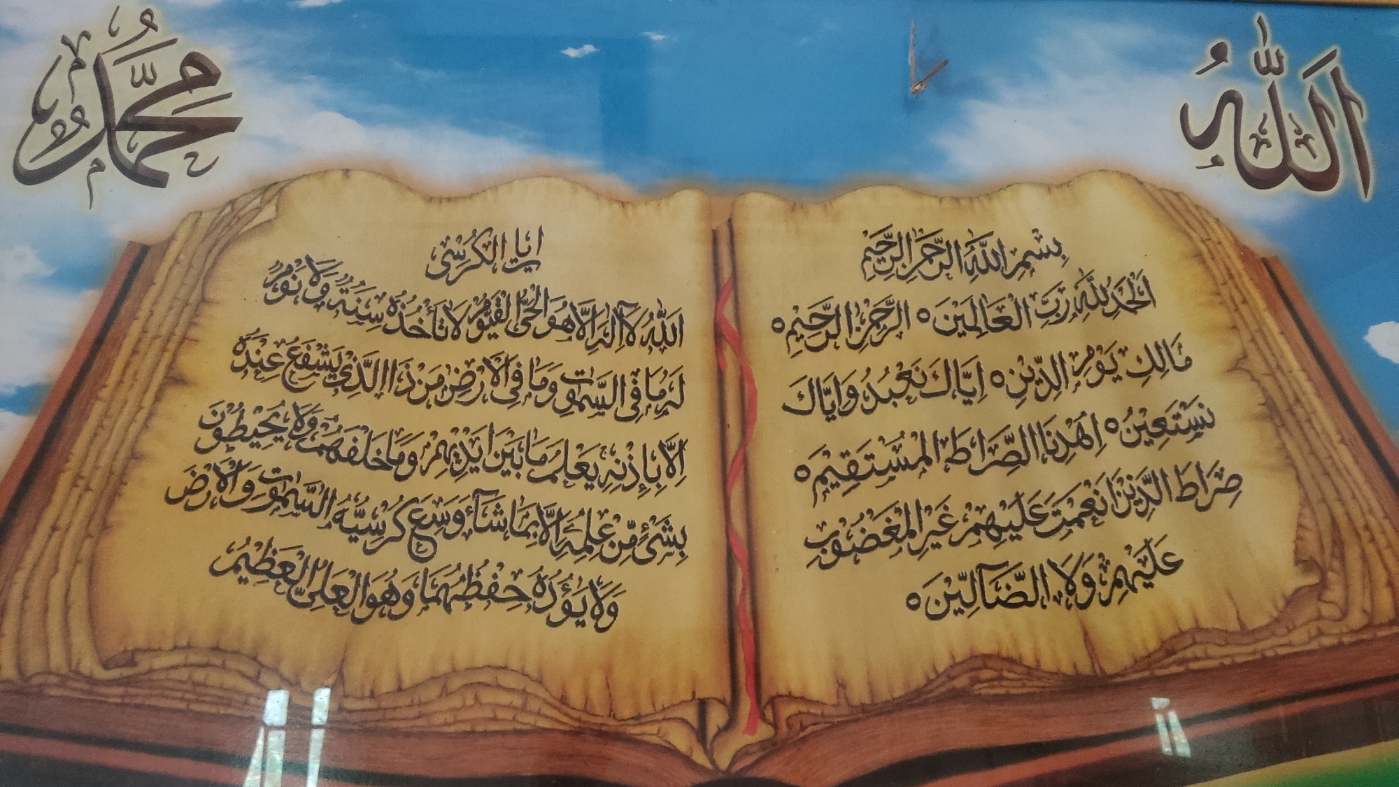 This image depicts an open book with Arabic calligraphy. The calligraphy appears to be from the Quran. On the left page, the text starts with "ان الكرسي" and continues downwards. On the right page, the text begins with "بسم الله الرحمن الرحيم" and proceeds with verses of Surah Al-Fatihah. Above the book, on the left, is a stylized Arabic word that could be "Muhammad" (محمد), and on the top right, the Arabic word for "Allah" (الله). The book is depicted as aged and weathered, with textured paper. The background is a blue sky with white clouds, suggesting an outdoor setting. The image is a decorative piece, likely a framed artwork or print, rather than a photograph of a real event or location. The information about "Ngadiharjo, Indonesia" is not visually evident in the image itself, as there are no geographical markers, buildings, or people identifiable as belonging to that specific location. Therefore, any description of this location cannot be derived from the provided image.