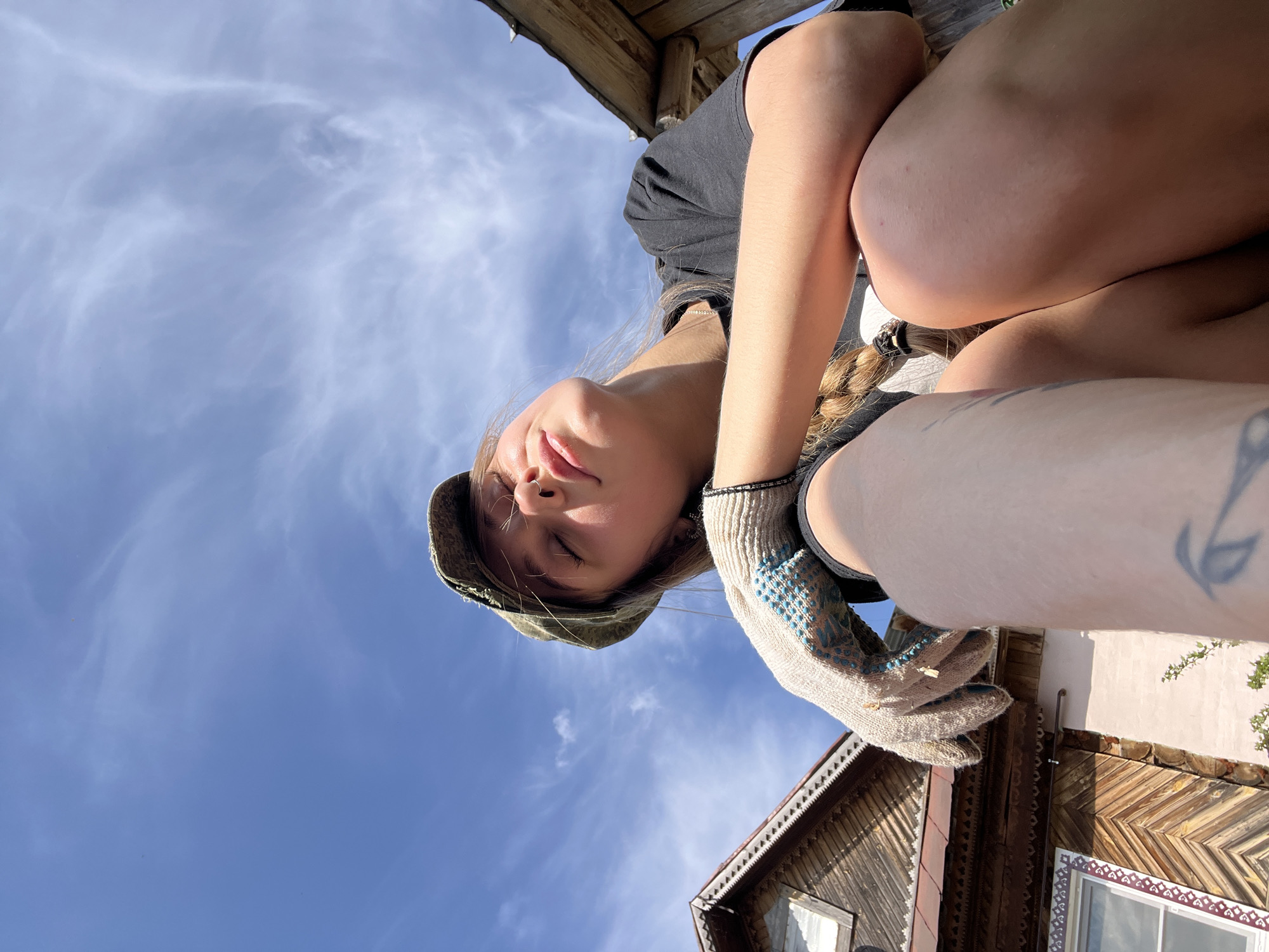 A low-angle shot depicts a young woman with light skin, closed eyes, and a relaxed expression. She wears a dark t-shirt and a military-style cap. Her blonde hair is braided and falls over her shoulder. She's wearing a work glove with blue patterned stitching on her right hand. A tattoo is visible on her arm.
Above her, the clear blue sky is lightly streaked with wispy clouds. Sections of a wooden building's roof and trim frame the image. The building has patterned wooden siding. The location may be the city of Фоки, Russia, based on the prompt. The photograph appears to have been taken from a slightly lower elevation, looking upwards toward the subject.