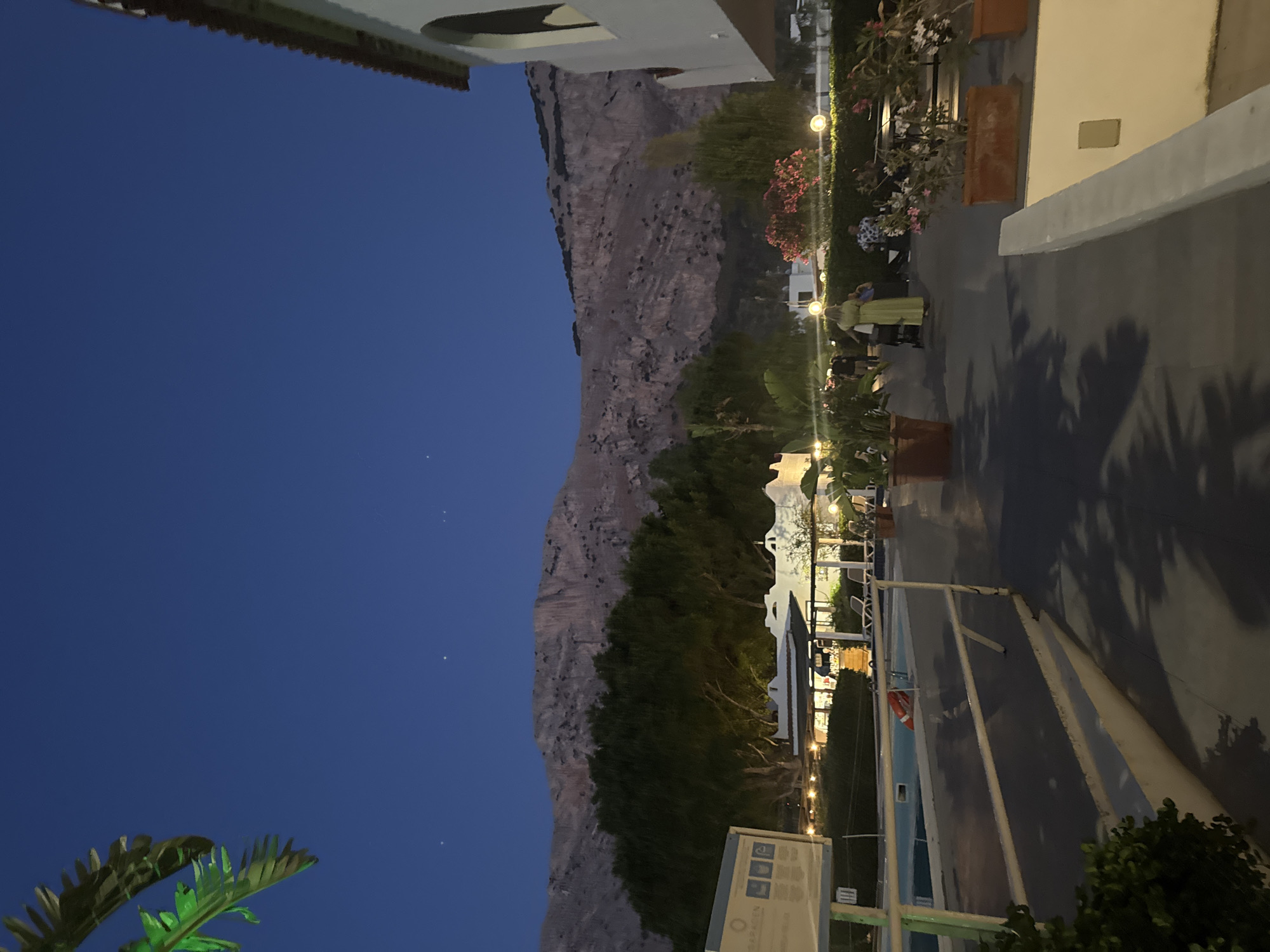 The image depicts a twilight scene at a resort or hotel near Isola delle Femmine, Italy. In the foreground, a paved walkway is visible, with a white railing to the left. Beyond the railing, a swimming pool can be seen, with a red life preserver floating in it. In the mid-ground, several people are walking and socializing. Some are pushing strollers. There are also potted plants and outdoor seating areas. In the background, lush green trees form a dense line, with a large, rocky mountain range rising behind them. The sky is a deep, clear blue, with a few faint stars visible. To the right, a white building with a tiled roof is partially visible, suggesting the presence of accommodations. A signpost with information in multiple languages is also visible to the left. The overall atmosphere is calm and relaxed, indicative of a vacation setting.