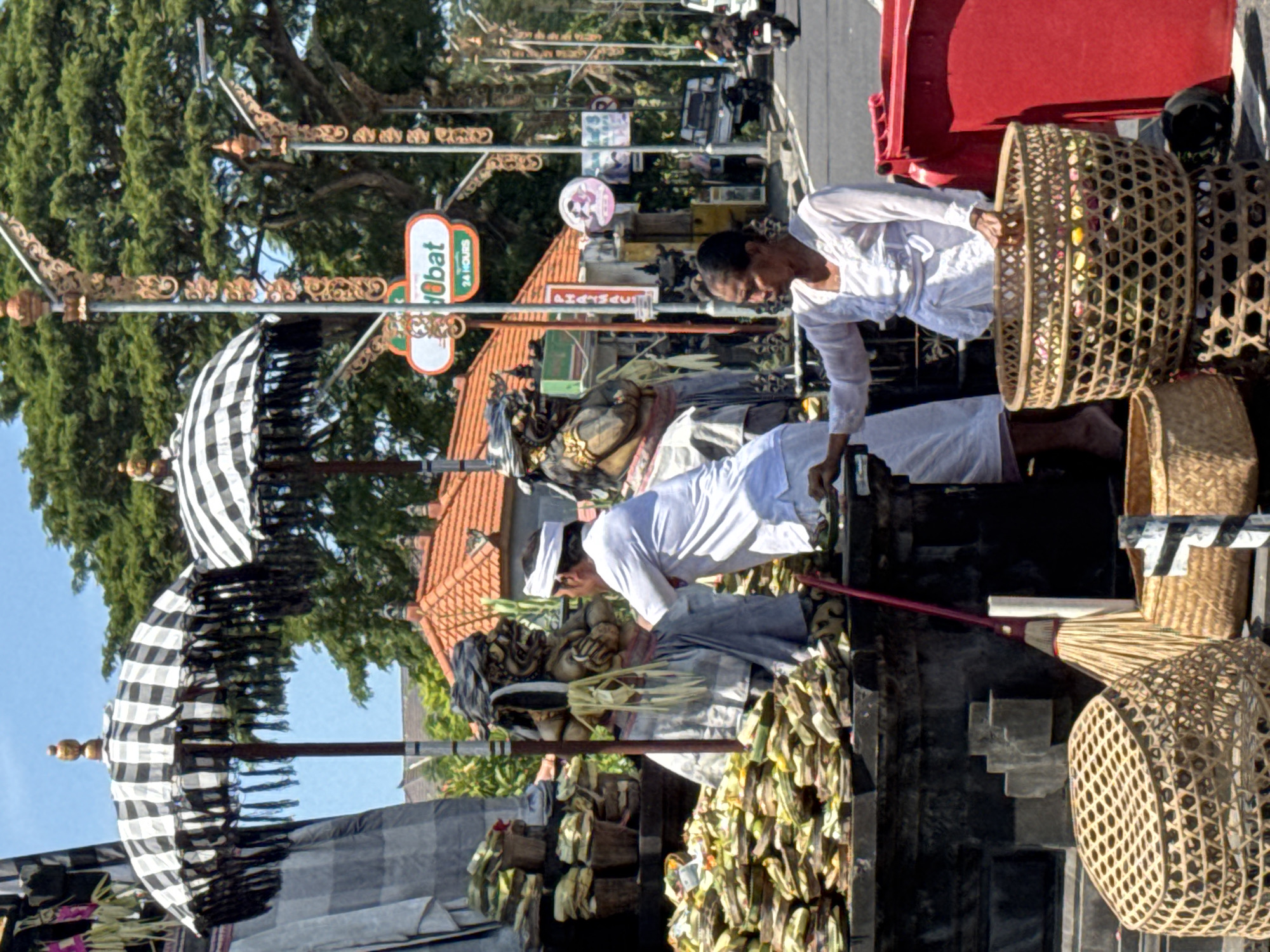 Captured in Nusa Dua, Indonesia, the image showcases two individuals participating in a religious ritual at what appears to be a temple or shrine. Both individuals, dressed in white ceremonial attire, are engaged in placing offerings. One person is kneeling, while the other is leaning towards the offerings, with woven baskets nearby. The temple or shrine is adorned with statues and numerous small, wrapped offerings. Black and white checkered ceremonial umbrellas flank the structure. The backdrop features a street scene, including trees, buildings with terracotta roofs, and street signs.