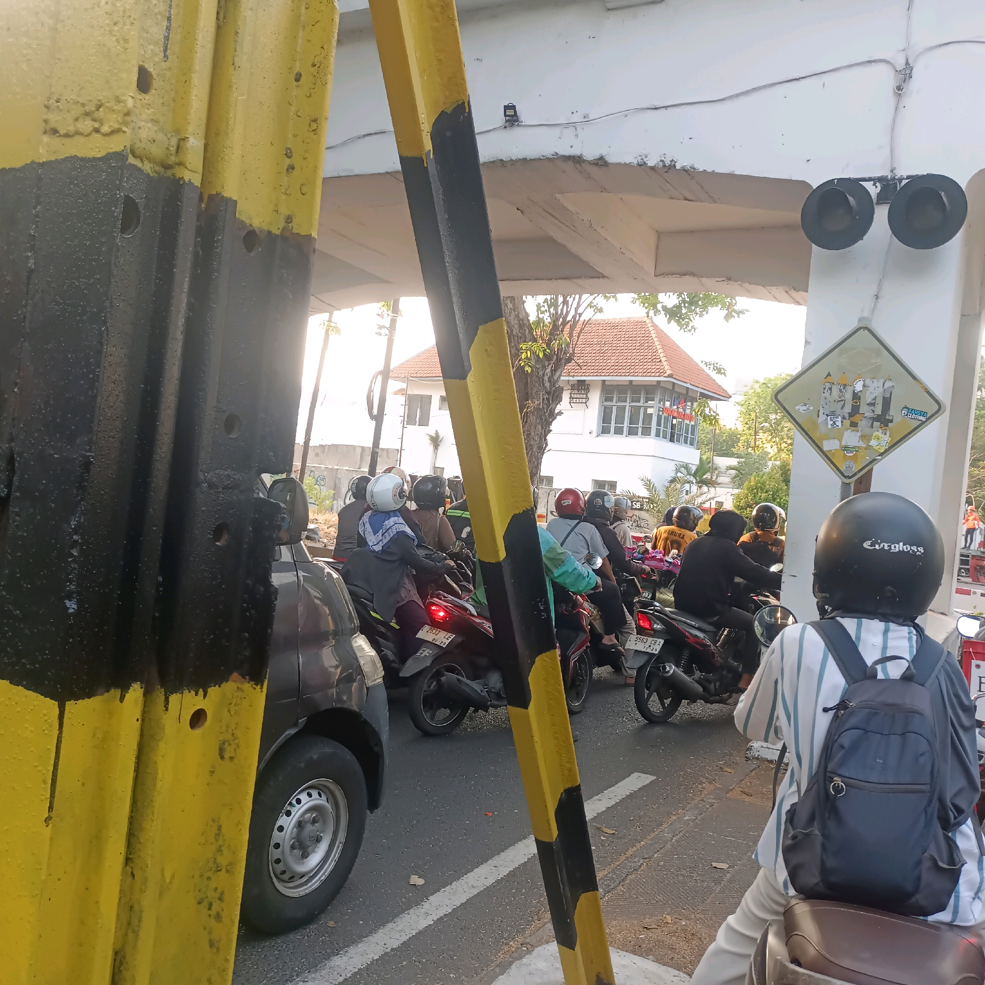 In Surabaya, Indonesia, a traffic jam is depicted at what appears to be a railway crossing. Prominently in the foreground, a large, yellow and black striped pole stands as a visual obstruction. To its right, a white overpass structure is visible, with railway signals – two dark, circular lights – mounted beneath it. Below the overpass, a building with a red tiled roof and white walls, possibly a signal box or station, is situated.
The road is filled with numerous motorcycles, suggesting a busy urban environment. Several individuals are seen riding these motorcycles, many wearing helmets in various colors, including white, black, and red. One person in the foreground is wearing a striped shirt and a black backpack. The vehicles are stopped, indicating a halt in traffic, likely due to an approaching train. The general lighting suggests it is daytime, possibly late afternoon given the warm tones. The scene is a snapshot of typical urban traffic in Surabaya.