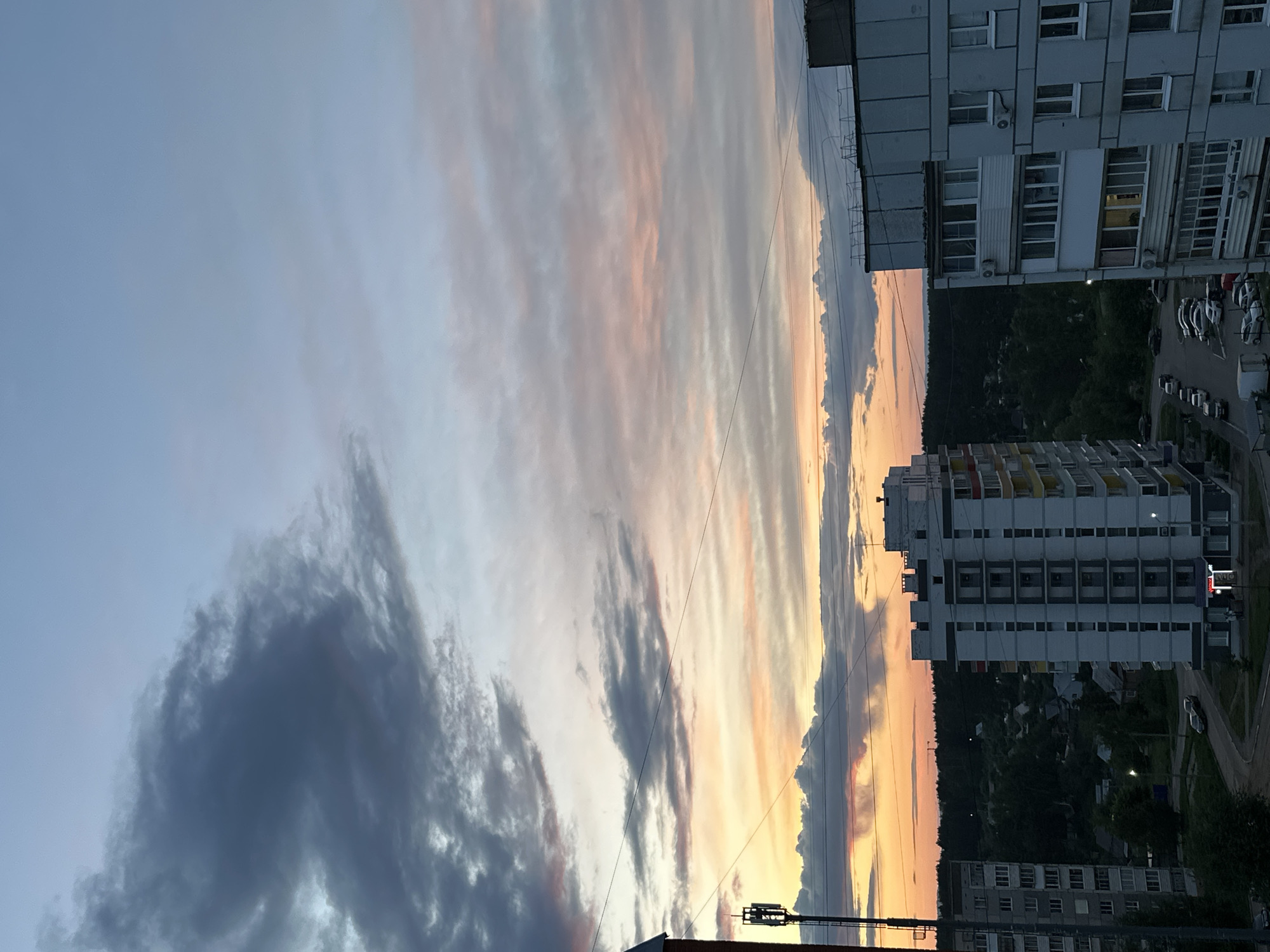 The image depicts a sunset sky over the city of Chaikovsky, Russia. The sky is filled with long, striated clouds, colored with shades of gray, orange, and pink, indicating the setting sun. Several apartment buildings are visible on the right side of the frame, typical of Soviet-era construction. A wooded area lies behind the buildings, and a parking lot with cars can be seen. Power lines intersect the sky in the foreground.