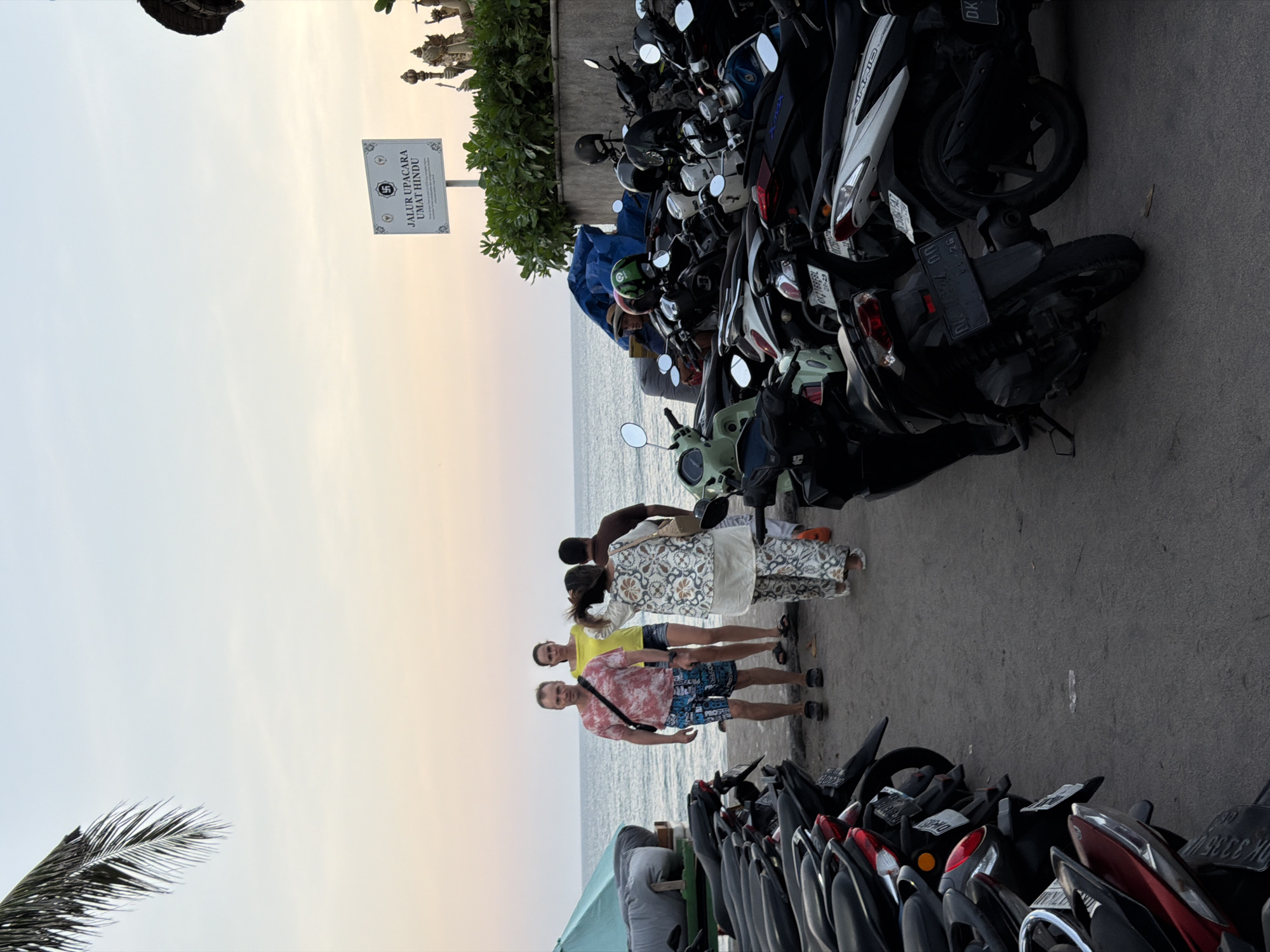 The image depicts a group of four people standing on a paved walkway beside a row of parked motorcycles. The people are facing the camera and appear to be posing for a photo. The foreground is dominated by the motorcycles, with a glimpse of the ocean in the background. The sky is overcast and has a soft, diffused light, suggesting it is either early morning or late afternoon. The location appears to be coastal, with the sea visible just beyond the walkway. There is a sign visible in the background that reads "JALUR UPACARA UMAT HINDU," which translates to "Hindu Community Ceremony Route." The overall mood is relaxed and casual, with the group enjoying their time together by the sea. The presence of numerous motorcycles suggests a popular tourist area or a local gathering spot.
