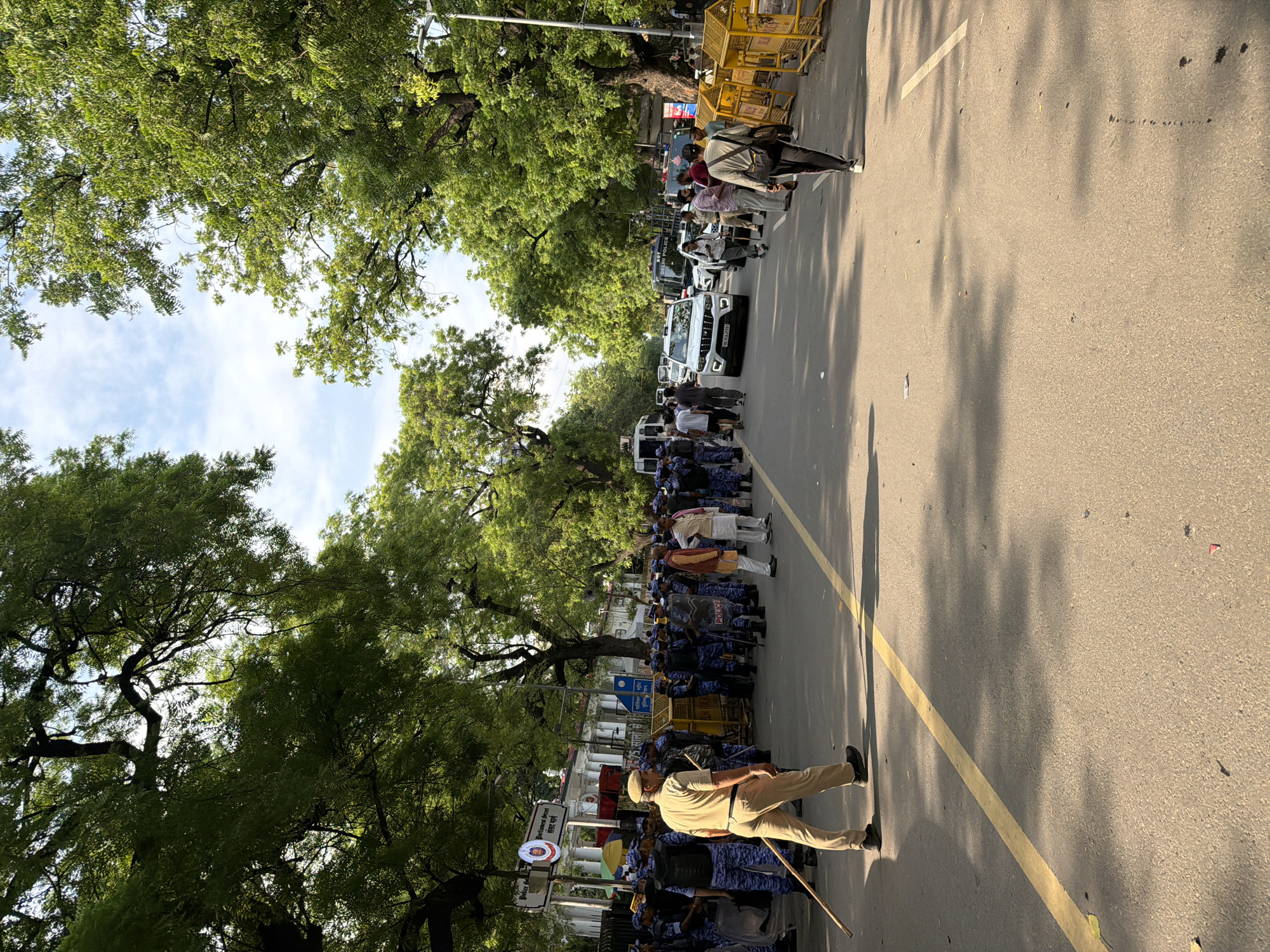 The image captures a street scene in New Delhi, India, likely near Parliament Street, indicated by a sign reading "Parliament Street" and "संसद मार्ग." The street is lined with mature trees providing shade, and the sky above is partially cloudy. A significant police presence is visible, with officers in blue uniforms and riot gear forming a line. One police officer in a tan uniform and cap, holding a stick, walks towards the viewer. In the background, several vehicles are present, including a white SUV and a Delhi Police van. Yellow barricades are set up along the side of the road. A group of civilians, some appearing to be protesters or onlookers, are also present, standing behind the police line. The overall atmosphere suggests a controlled environment, possibly due to a protest or demonstration.