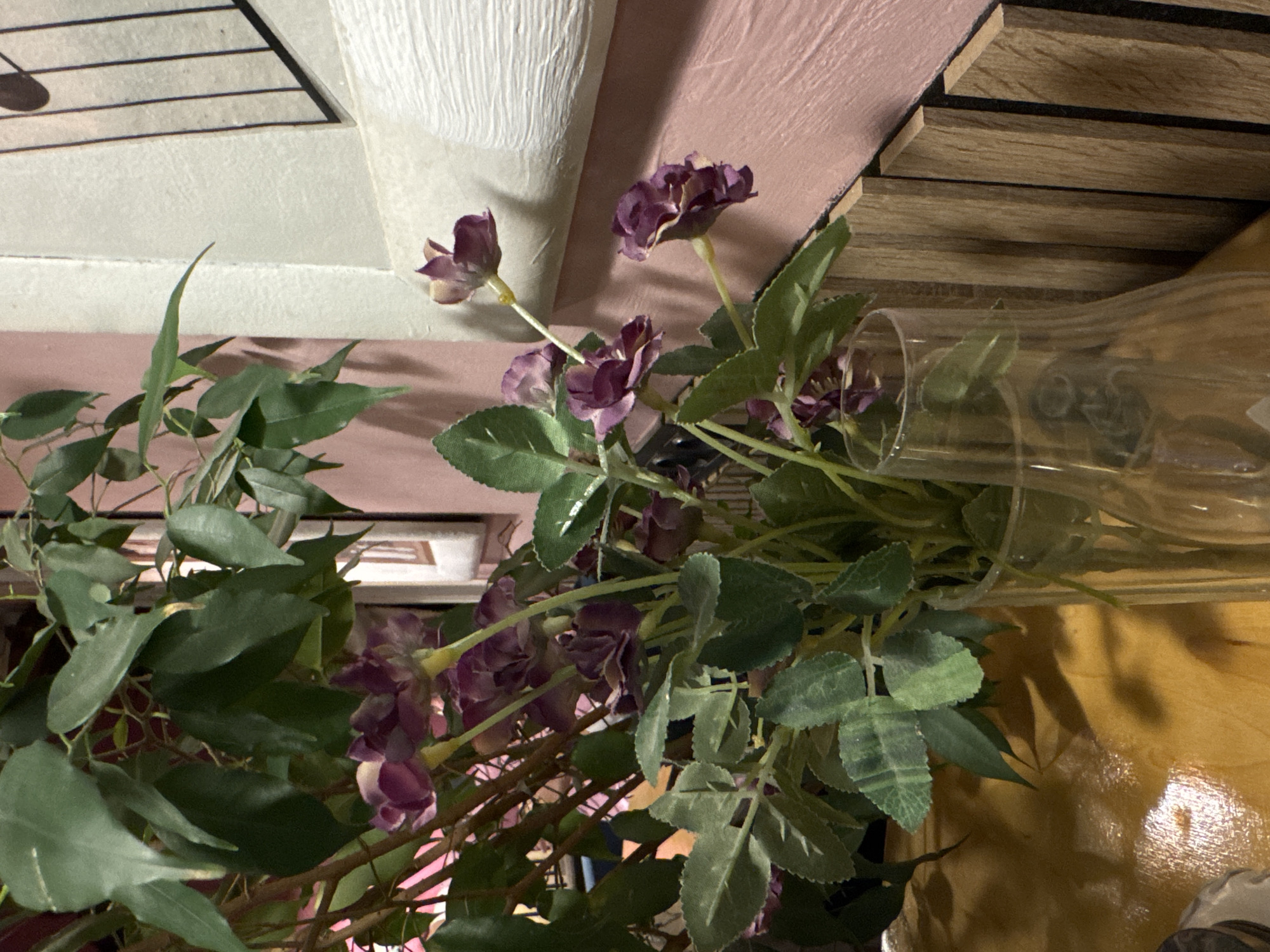 In Minsk, Belarus, this image captures a close-up view of a bouquet of artificial purple flowers and green foliage in a clear glass vase. The vase sits on a glossy wooden surface, possibly a table. Behind the flowers, a textured pale pink wall is visible, adorned with a vertical decorative element featuring thin black lines and a curved outline. To the right of the wall, a white textured surface with horizontal ridges can be seen. The overall lighting is dim, suggesting an indoor setting, possibly a cafe, restaurant, or private residence in Minsk.