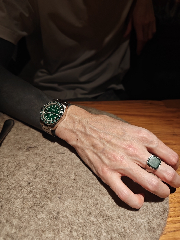 A person’s hand rests on a rough, gray surface with a dark wood table visible. The arm, seemingly tattooed black, wears a silver wristwatch with a green face. On the ring finger is a silver ring with a dark green, possibly jade, stone. The person wears a gray shirt. The scene is dimly lit.