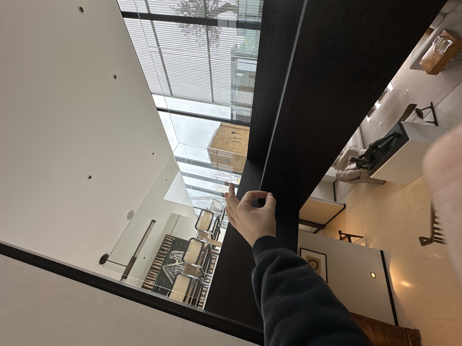 The image depicts a multi-level interior of a modern building, likely a high-end private residence or gallery, located in London, United Kingdom. The perspective is from an elevated position looking downwards and across.
In the immediate foreground, a left hand, wearing a dark long-sleeved garment, performs an "okay" gesture just above a dark brown, wide balustrade or railing that diagonally traverses the lower left to central frame.
Beyond the balustrade, an upper level is visible, separated by a clear glass railing. This level contains a dining area featuring a long, light-colored wooden table with four chairs characterized by metal frames and cane backs. A rectangular, dark pendant light fixture hangs above the table. On the wall behind the dining area, a large tapestry or artwork with an abstract, possibly avian, design is displayed.
Through expansive floor-to-ceiling windows on the far side of the upper level, an exterior view reveals vertical architectural slats or louvers and the upper branches of a tree. A large, light-colored wooden shipping crate, marked with "FRAGILE" and upward-pointing arrows, is visible through these windows, positioned outdoors.
Below the viewpoint and the upper level, a spacious lower floor area is visible, featuring light-colored tiled flooring. A bronze reclining female sculpture rests on a prominent white pedestal. Portions of light-colored sofas, a glass-topped coffee table, and other modern furnishings are present. The walls are white with recessed lighting fixtures, and a framed artwork is partially visible on a wall to the left. The scene indicates a carefully curated, well-lit environment with a focus on contemporary design and art.
