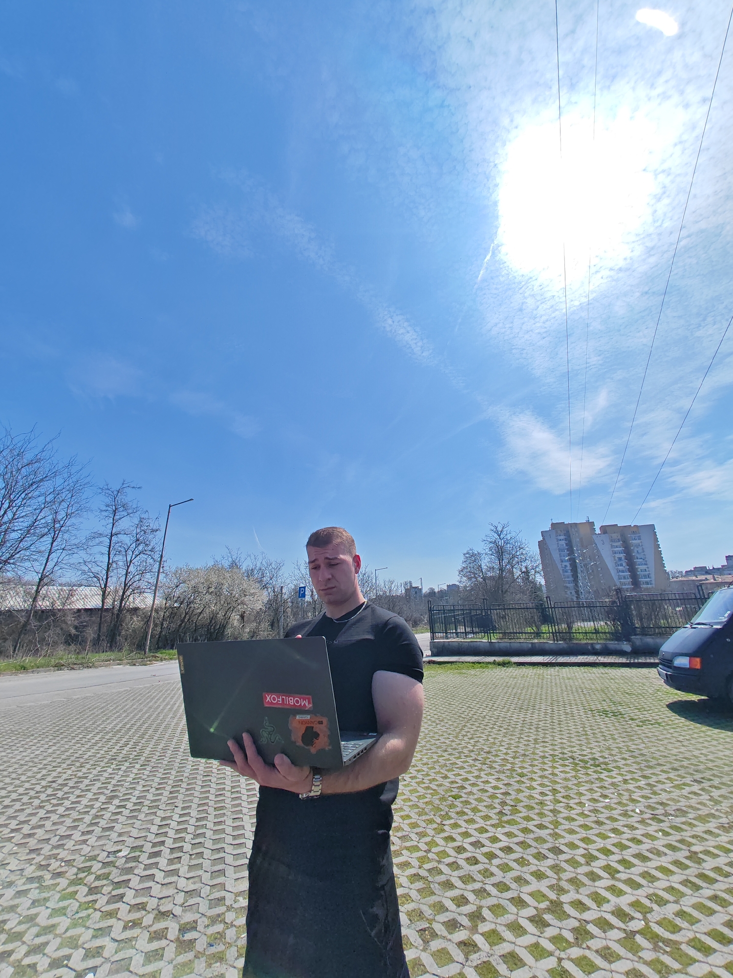 A male individual, estimated to be in his 20s or 30s, stands outdoors in Shumen, Bulgaria, holding a dark-colored laptop. He has short brown hair, is wearing a black short-sleeved t-shirt and dark trousers, and a silver-colored chain around his neck. His arms display visible musculature. He is looking down at the open laptop screen, which he holds with both hands. The back of the laptop lid features multiple stickers, including a red one with "MOBILFOX" in white text, a black one with a green Razer logo, and another orange-brownish sticker with "GARON GAMING."
The individual is standing on a patterned grey concrete paver surface with grass growing in the gaps, indicative of a parking area or pedestrian zone. To the left, an asphalt road is visible, bordered by bare deciduous trees and some trees with white blossoms, suggesting springtime. A street light pole is also present on the left. To the right and behind the individual, a dark-colored van is partially visible, parked on the paved surface. Beyond a dark metal fence, a multi-story residential building, consistent with Eastern European "panelka" architecture, stands prominently, featuring yellow and white facade elements and the number "3" on its side. The sky above is clear blue with scattered white clouds, and the sun is brightly visible in the upper right quadrant, creating a strong glare. Several thin power lines traverse the upper sky.