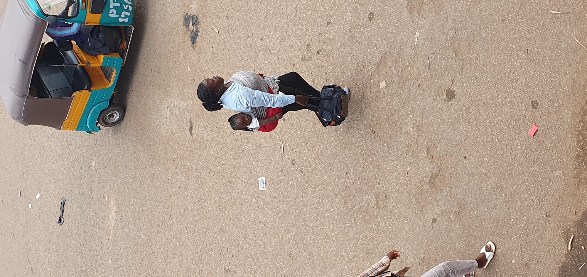 In Bukuru, Nigeria, a woman walks across a dusty, tan-colored road, carrying a baby on her back. The baby is wrapped in a red cloth and a grey sling, with its head resting on the woman's shoulder. The woman wears a light blue shirt and black leggings, and carries a dark blue duffel bag in her right hand. A three-wheeled auto-rickshaw, painted turquoise and yellow, is parked in the upper right portion of the frame, with a person in a blue garment seated in the driver's seat. Other scattered debris is visible on the road surface, including white paper scraps and dark, indeterminate objects. In the lower left, a person wearing light-colored pants and white sandals is partially visible.