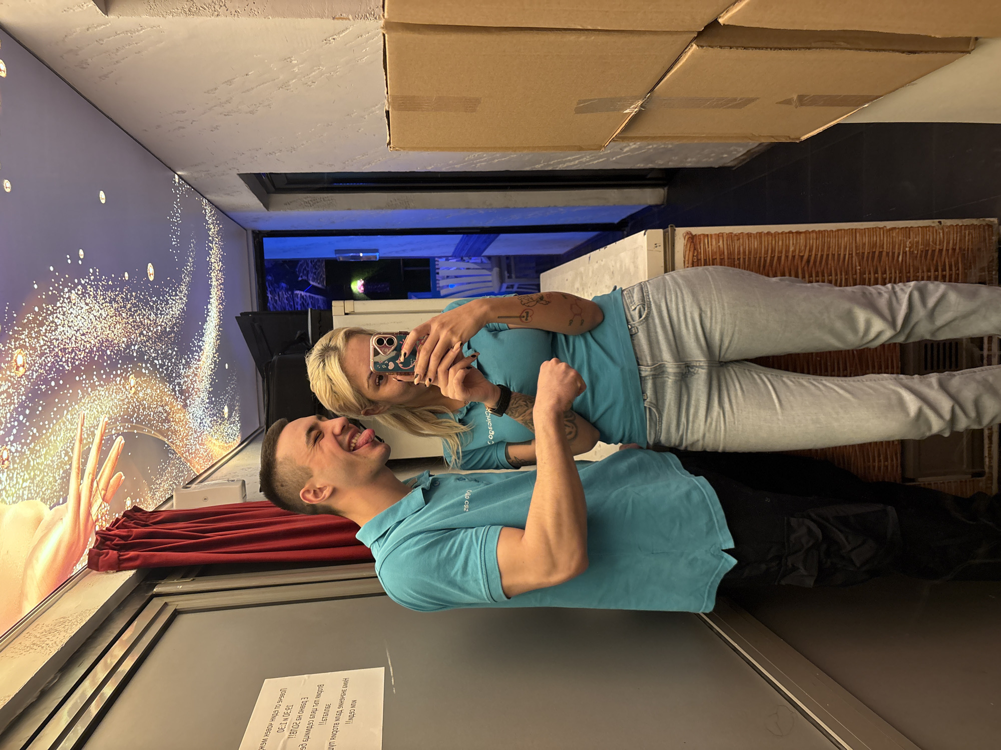The image, when correctly oriented, depicts two individuals, a woman and a man, taking a playful selfie or video indoors. The woman, with blonde hair, wears a light blue polo shirt, light blue jeans, and has a tattoo on her right arm. She holds a smartphone with a distinctive case featuring large eyes, aiming it towards herself and the man. She smiles subtly at the phone. Beside her, the man, with short dark hair, wears a matching light blue polo shirt and black pants. He looks towards the camera with a wide, open-mouthed smile and his tongue playfully sticking out. Both shirts feature a partially visible logo, possibly related to "Copacabana" or a similar brand, hinting at uniforms.
The setting appears to be a commercial or entertainment space, possibly an interactive attraction or photobooth. To their left, a large, glowing abstract hand and sparkling light effects are projected onto a curved wall, partially obscured by a gathered red curtain, suggesting a thematic or magical ambiance. On the right, textured brown paneling is visible, with cardboard storage boxes stacked on shelves above. A dark floor and a distant blue-lit area complete the background.
A white sign, attached to a surface in the lower left foreground, contains Bulgarian text. It reads: "ВНИМАНИЕ ЗОНИ СВЕТИЛНИ!" (Attention, lighting zones!), "НИЕ СВЕТИМ!!" (We light up!!), "ВХОДОВЕТЕ СТАРТИРАТ СЛЕДНАТА ПРОГРАМА:" (Entrances start the following program:), followed by time slots "18:30 и 20:00 ч." (18:30 and 20:00 h). Further text mentions "ОБОРУДВАНЕ ЗА СНИМКИ" (Equipment for photos), "Влизате в свят на магия и приключения!" (You enter a world of magic and adventure!), and a date "21.08.23". Additional entrance times are listed as "10:00 - 12:00 ч." and "16:30 - 18:30 ч.". The presence of Bulgarian text confirms the location context of Plovdiv, Bulgaria. The subjects' joyful expressions and the interactive environment suggest a fun, casual activity.