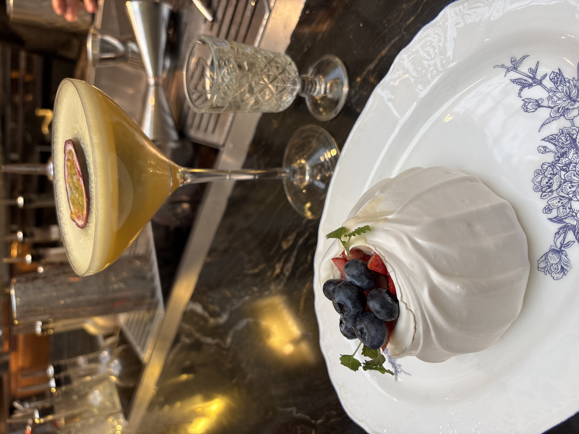 The image presents a close-up, slightly overhead view of a sophisticated dessert and cocktail arrangement on a dark, reflective surface, likely a bar counter or restaurant table, in Saint Petersburg, Russia.
In the foreground, a vibrant yellow cocktail, possibly a passion fruit martini, is served in a classic stemmed martini glass. The drink features a frothy top and is elegantly garnished with a half-slice of fresh passion fruit, showcasing its distinct seeds and pulp. Behind the cocktail, an empty, small, stemmed glass with a decorative cut-glass pattern is visible. The blurred background reveals other elements of a bar setting, including stainless steel bar tools like a jigger and shaker, and additional glassware, indicating an indoor venue such as a restaurant or bar.
To the right, a delicate pavlova dessert is placed on a round, white plate. The pavlova consists of a crisp, white meringue shell, artfully shaped into a cup and filled with an abundance of fresh, deep blue blueberries and bright red sliced strawberries. A small sprig of green herb, likely mint or lemon balm, garnishes the fruit filling. The white plate itself features an elegant blue floral pattern along one side of its rim, enhancing the presentation of the dessert.
The lighting is soft and ambient, suggesting an indoor evening setting typical for dining or drinks. The dark, glossy table surface reflects parts of the drinks and plates, adding depth to the scene. No people are visible, and there is no discernible text in the image. The overall composition conveys a sense of refined indulgence.