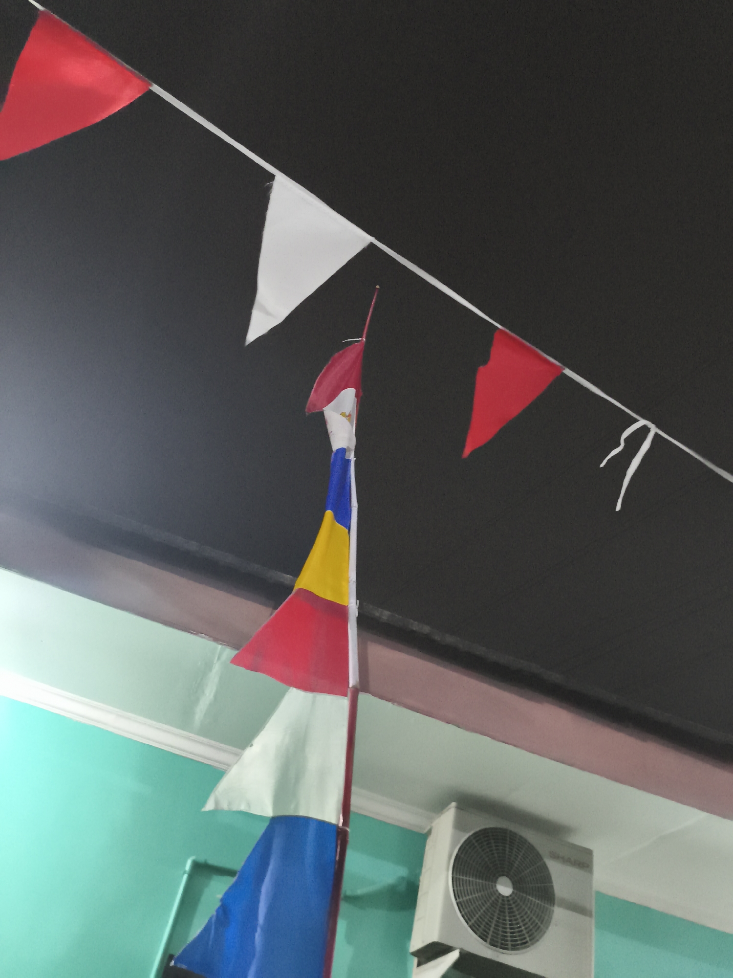 The image shows a string of triangular flags, some red and white, others with multiple colors including red, yellow, and blue. The flags are strung against a dark background, likely a ceiling or overhang. Below the flags, a section of a teal-colored wall is visible, with a white decorative trim along the top. To the right, an air conditioning unit with the brand name "SHARP" is mounted on the wall. The scene appears to be outdoors or in a covered outdoor area, possibly a venue for an event or celebration, given the festive nature of the flags. The overall impression is one of decoration and possibly anticipation of an event. There are no people visible in the image. The image is taken from a low angle, looking upwards.