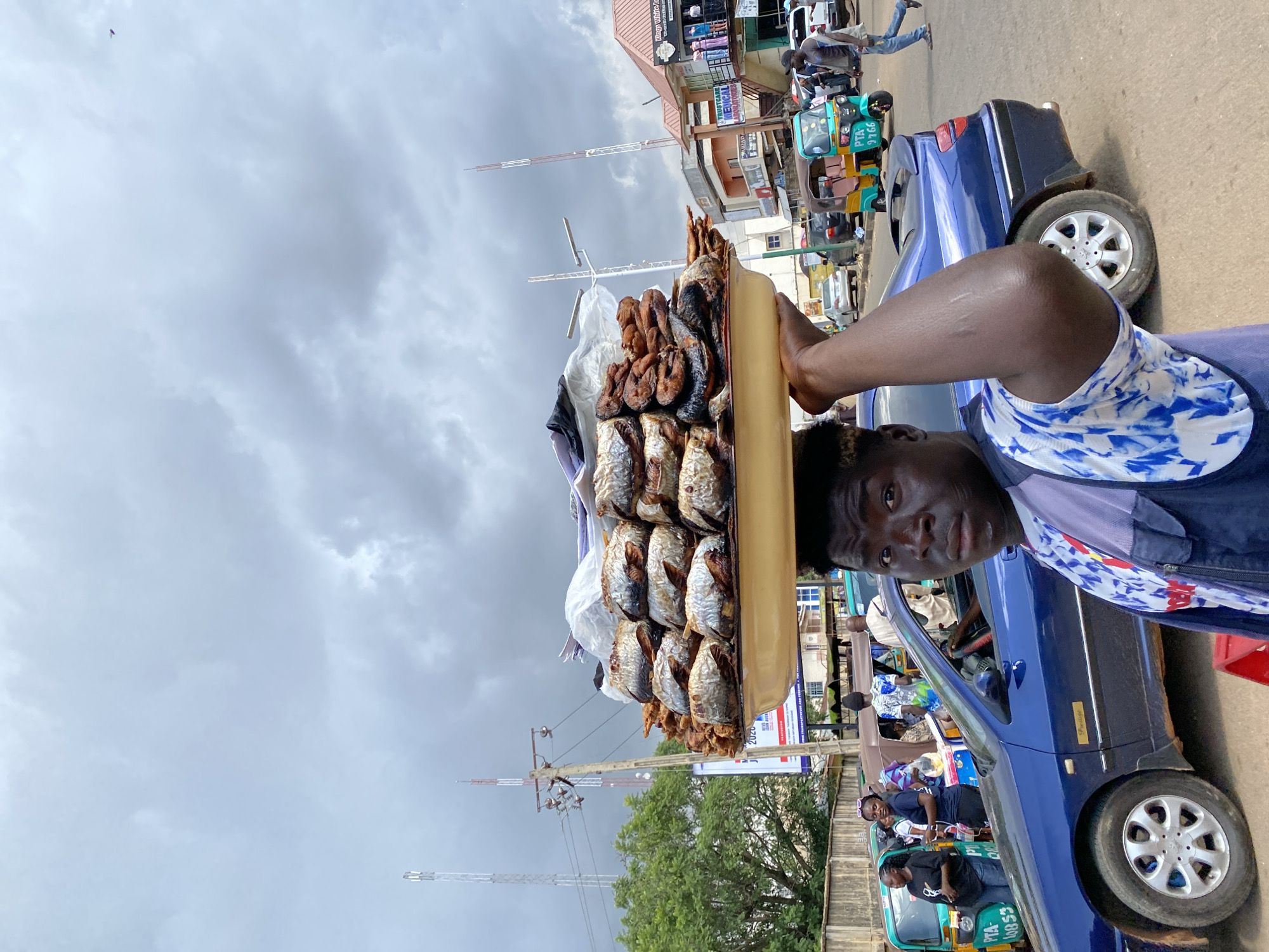 The image shows a street scene in Jos, Nigeria. The main subject is a person, likely a vendor, holding a large tray of dried fish. The tray is overflowing with fish, some larger and whole, others cut into pieces. The vendor is partially visible, wearing a blue and white patterned top and a dark blue jacket. Their face is looking towards the camera.
The scene is a busy street, indicated by the presence of a blue car, several auto-rickshaws (Keke Napep), and other vehicles in the background. People are visible both in and around the vehicles. Some are passengers, while others appear to be walking or standing by the roadside. The overall atmosphere suggests a market or roadside commercial area.
The weather appears to be overcast, with a cloudy sky dominating the upper portion of the image. This could imply it is either early morning or late afternoon, or simply a day with heavy cloud cover. The presence of tall communication towers and what looks like a building with signage suggests an urban or semi-urban environment.
Notable details include the branding on the Keke Napep, with text such as "PTA 9766" and "PTA 10853". One of the buildings in the background has a sign that is partially readable, including the words "TRUSTCARE MEDICAL". The image is oriented vertically, and the perspective is from a person standing or sitting near ground level, looking slightly upwards. The overall impression is that of a vibrant, everyday street scene in a Nigerian city.