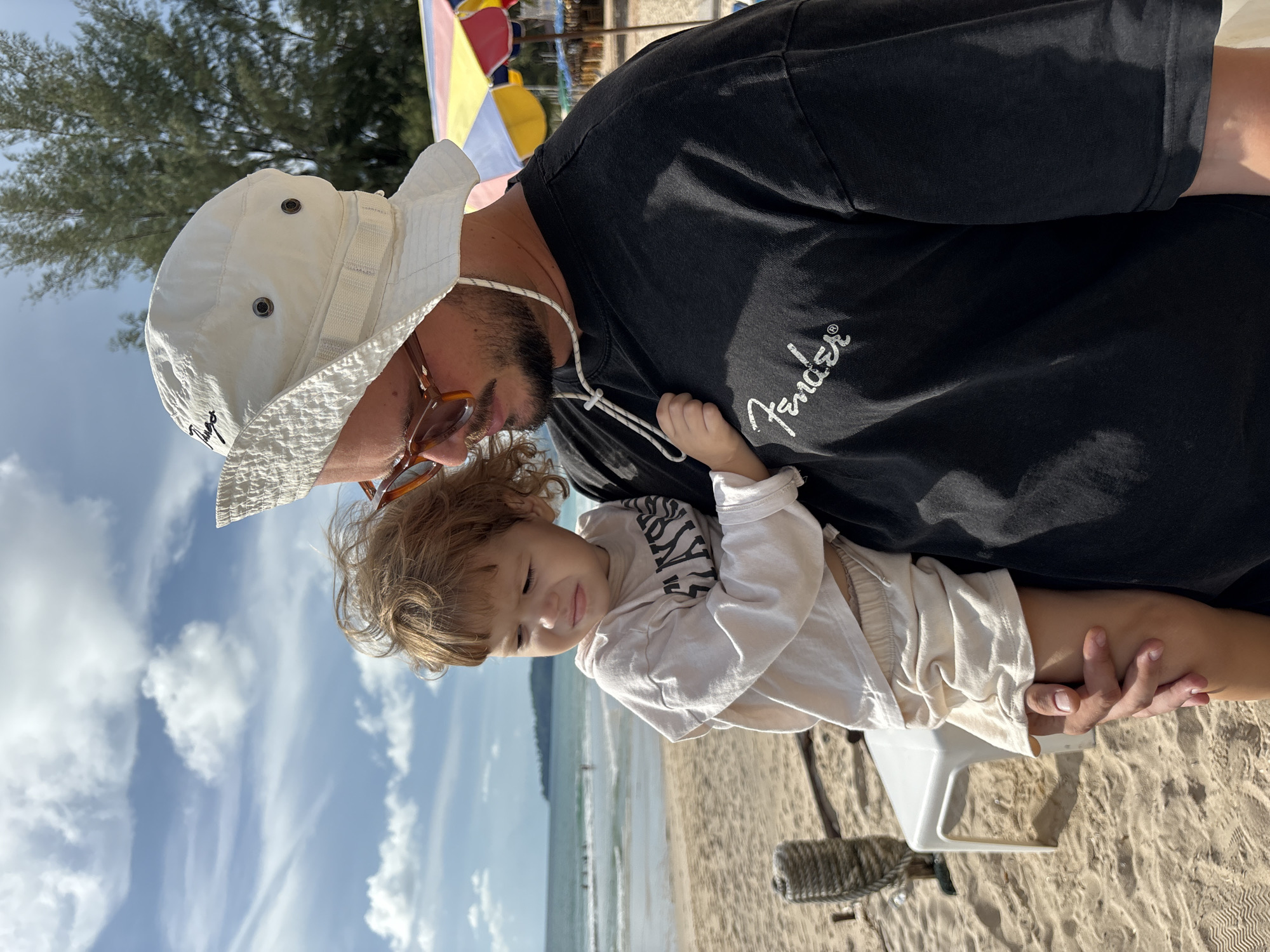 A man wearing a white bucket hat and sunglasses holds a young child on a sandy beach. The child, with light brown curly hair, is wearing a cream-colored long-sleeved shirt and shorts, and appears to be crying. The man is wearing a dark t-shirt with the word "Fender" printed on it in white script. In the background, the ocean with gentle waves is visible, along with a few beach umbrellas and trees. The setting is the beachfront area of Choeng Thale, Thailand.
