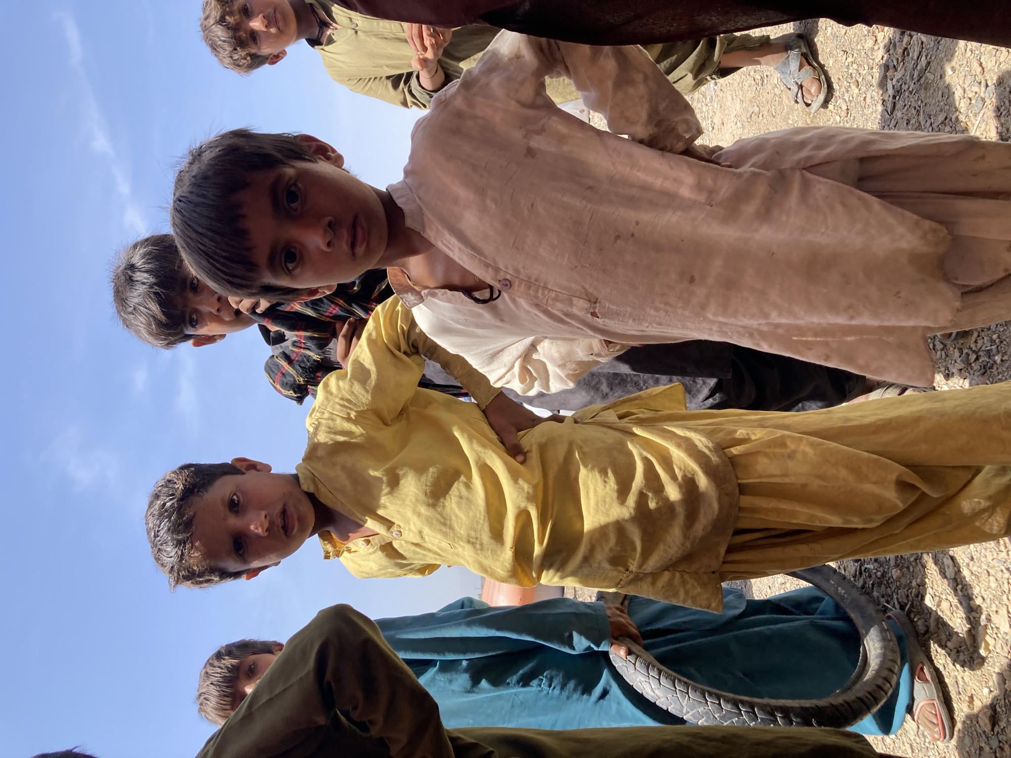 In Hussenāni, Pakistan, a group of boys wearing traditional South Asian clothing stand together. The image is taken from a low angle, looking upwards. One boy wears a yellow shalwar kameez, another a white one, and a third a blue one. A tire is visible in the lower right of the frame, being held by someone wearing the blue outfit. The ground is gravel. The sky is visible in the upper left.