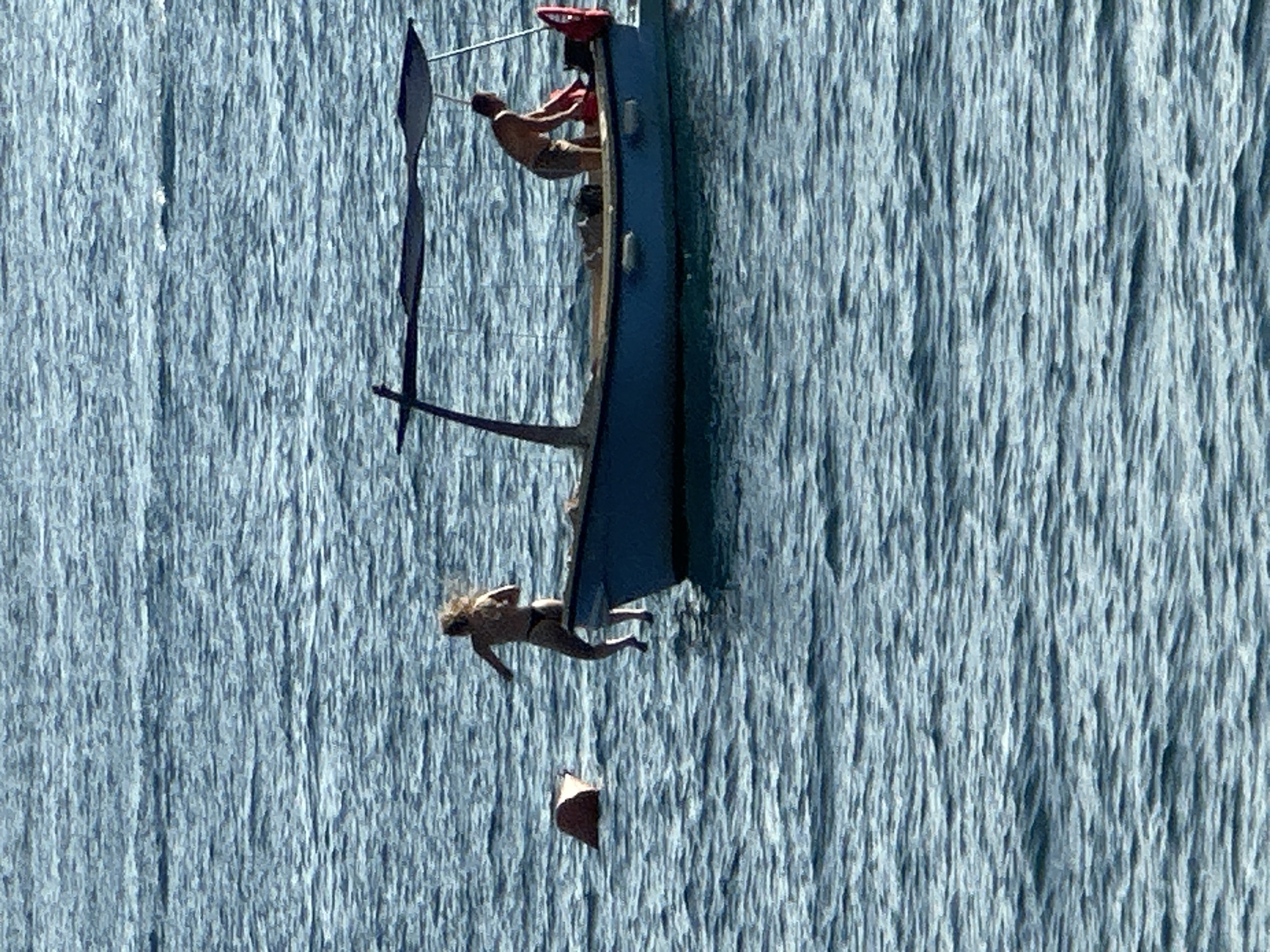 Two individuals are in the water next to a blue boat under a canopy. A woman in a bikini is in mid-air, jumping from the boat into the water, with her hair blowing behind her. A man wearing shorts is inside the boat, bending over to pick up a red bag. Another red object, possibly a buoy, is floating in the water to the left of the boat. The scene takes place on a bright, sunny day on the water, which appears to be the Adriatic Sea near Budva, Montenegro, known for its coastline and numerous boats.