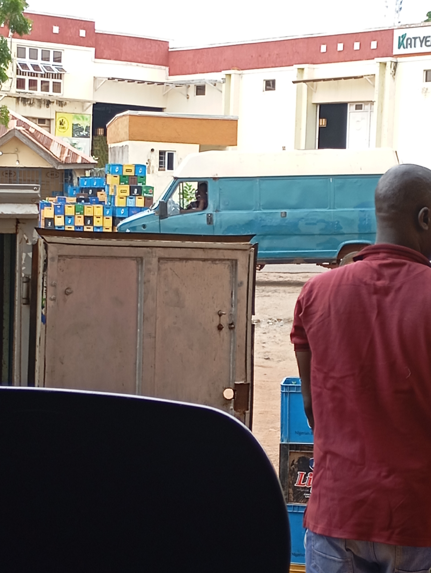 In Shendam, Nigeria, a scene unfolds at a distribution point. A man wearing a red shirt and blue jeans stands facing away from the camera, with his back to a large, open metal doorway. To his right, a turquoise van is parked, its side facing the viewer. Stacked high behind the van are numerous plastic crates filled with beverage bottles. The crates are a mix of yellow and blue. The background reveals a building with red and white facades, featuring rows of windows and a prominent green sign with yellow lettering. The ground appears to be dirt or gravel. The foreground is dominated by the dark silhouette of a vehicle's interior, framing the view of the outdoor scene.